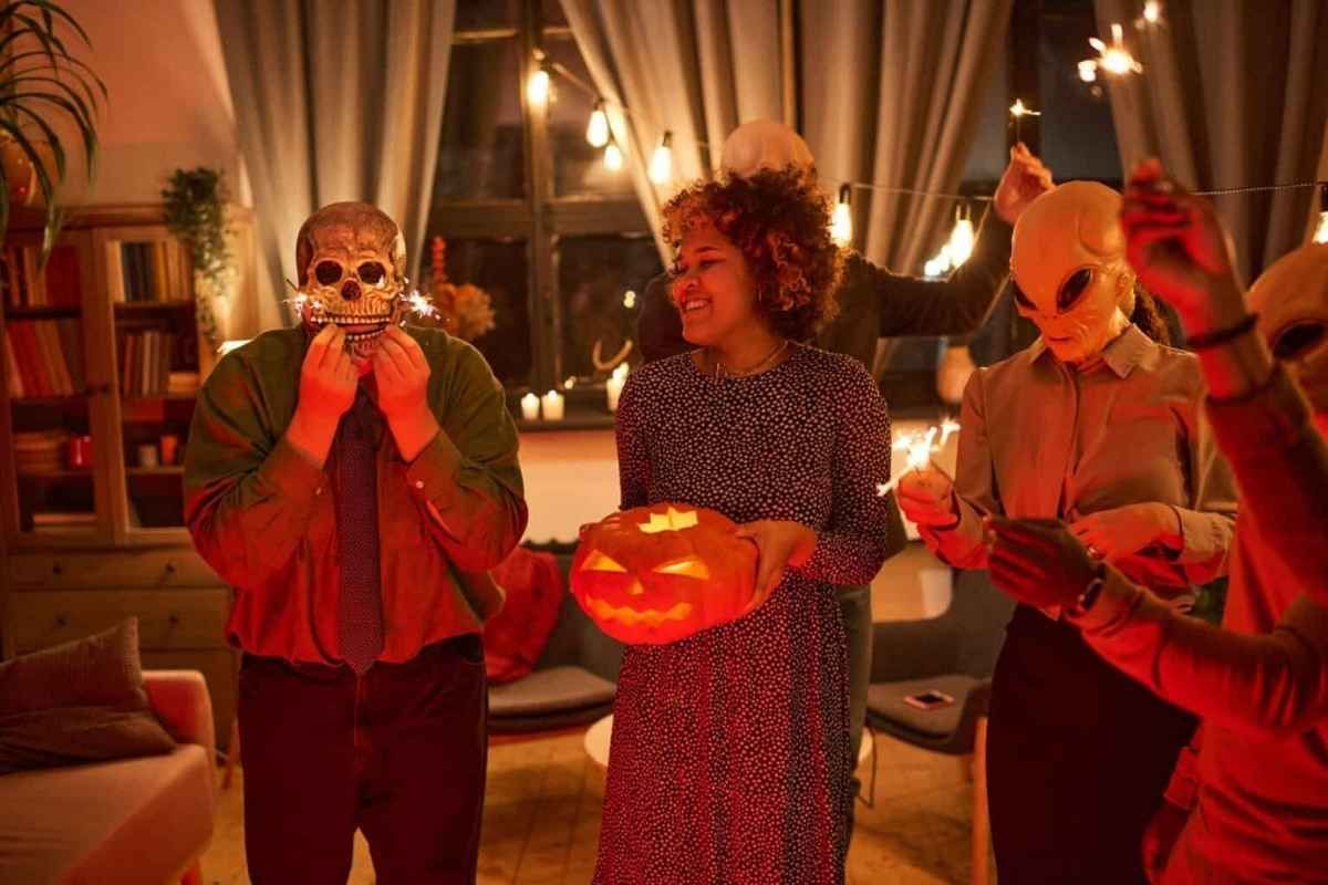 People in costumes at a Halloween party, holding a skull, a carved pumpkin, and sparklers in a warmly lit living room.