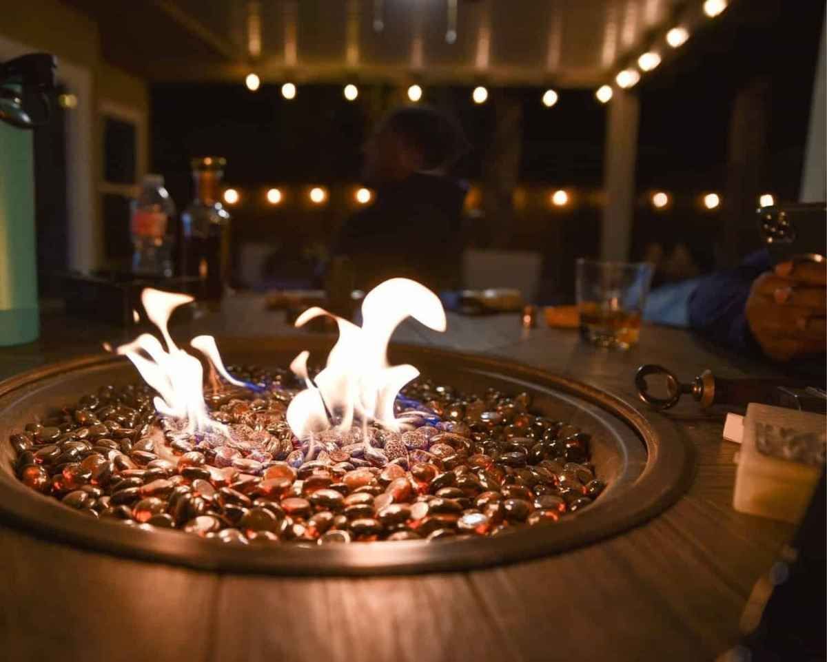 Fire pit with flames, surrounded by dark red glass rocks on a wooden table outdoors at night.
