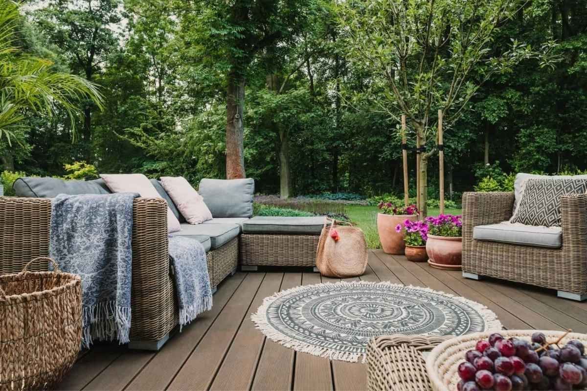 Outdoor patio with wicker furniture, rug, and potted plants in a lush green garden setting.