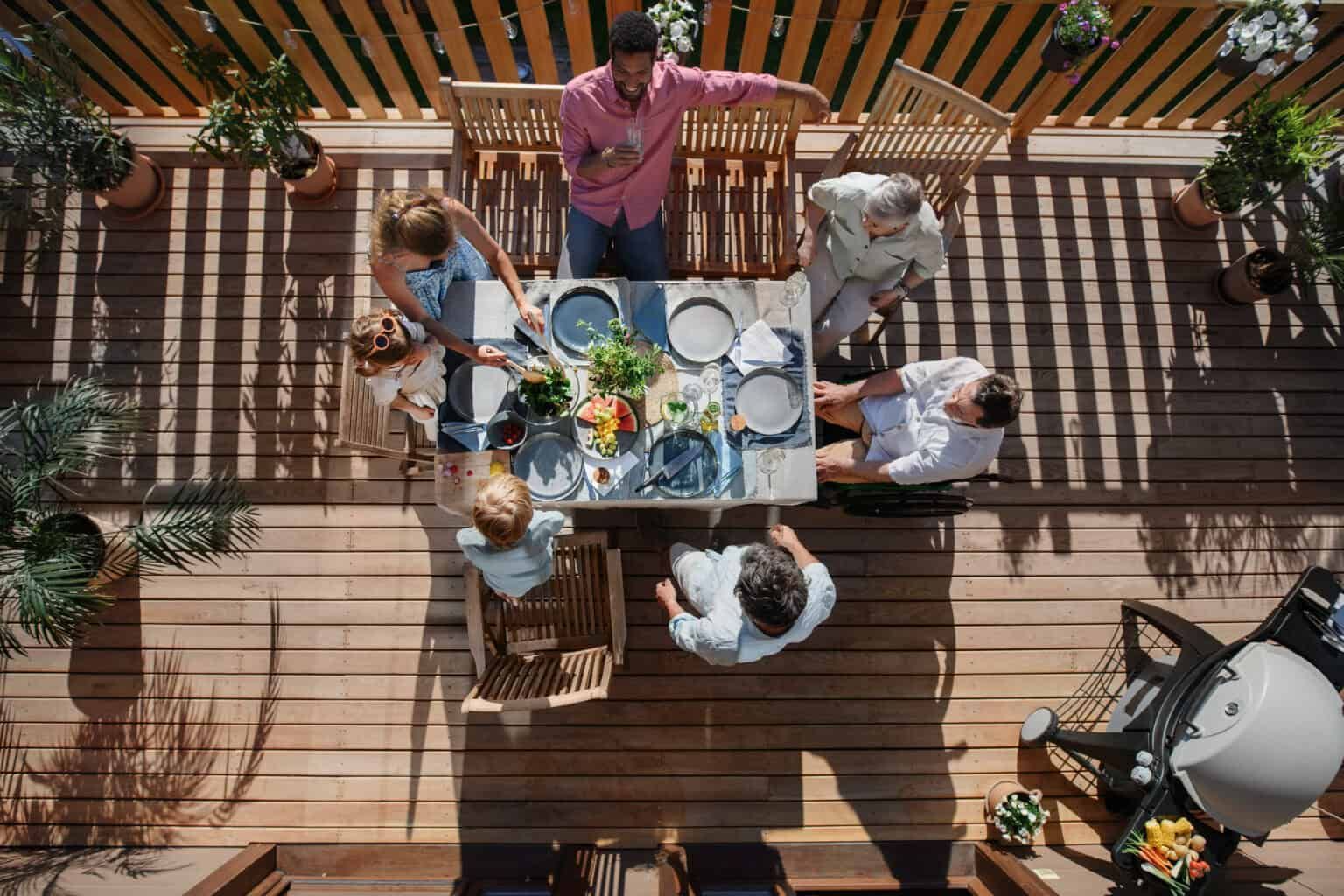 Family gathering around a table on a wooden deck, preparing a meal. Sunlight, greenery, and a grill are visible.