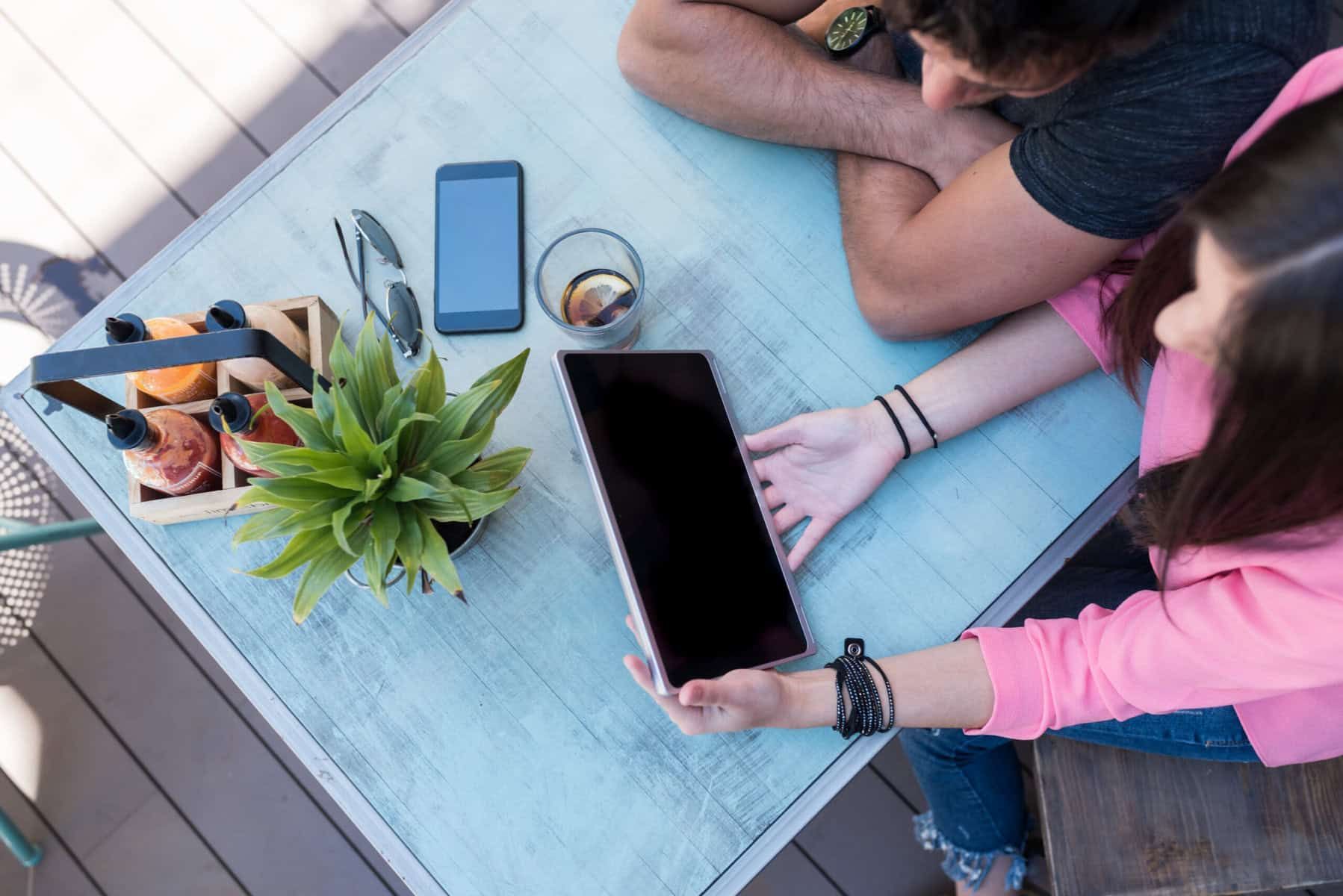 Two people at a table, looking at a tablet. A phone, plant, drinks, and sunglasses are also on the table.