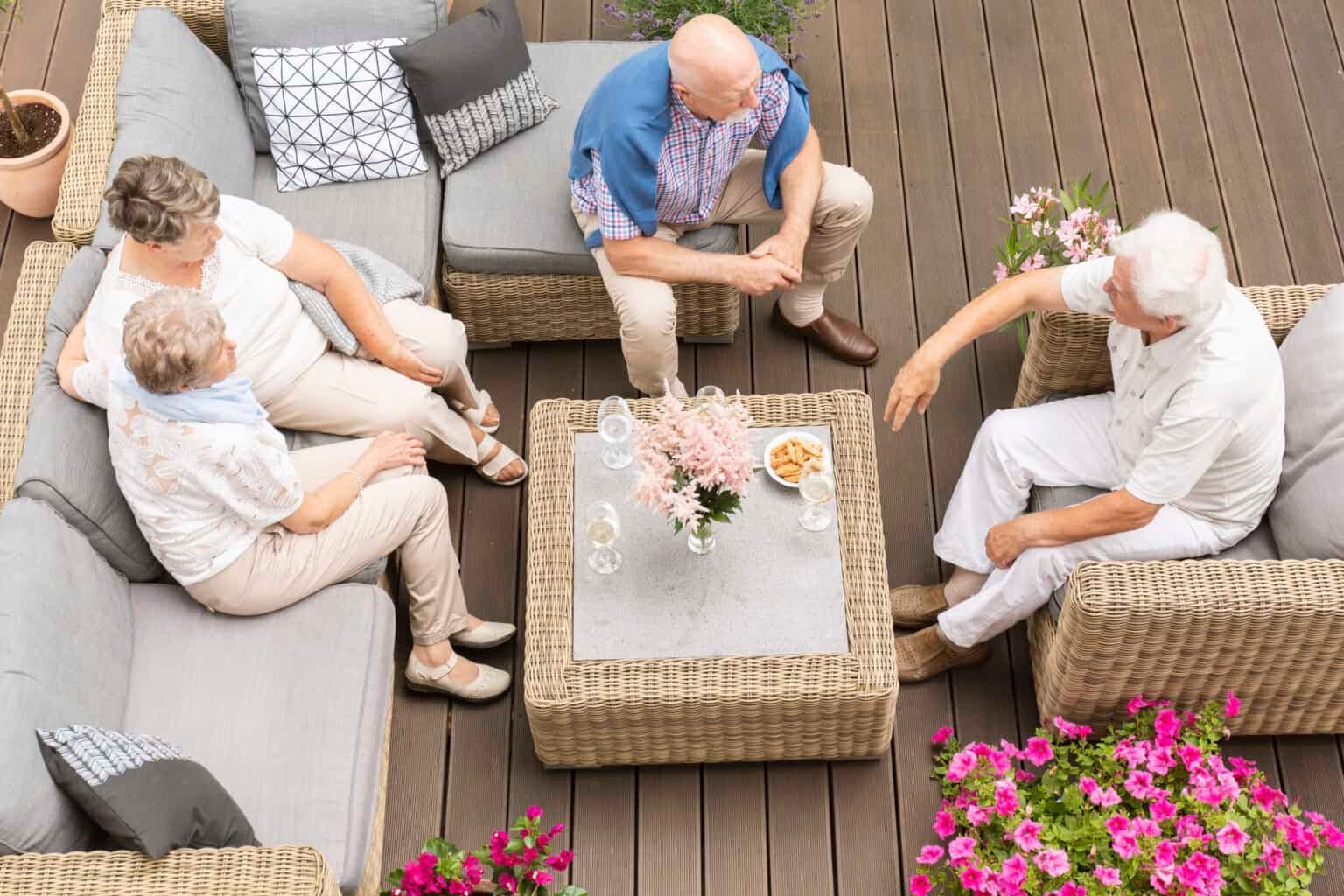 Group of people seated on outdoor wicker furniture, socializing around a table with snacks and flowers.