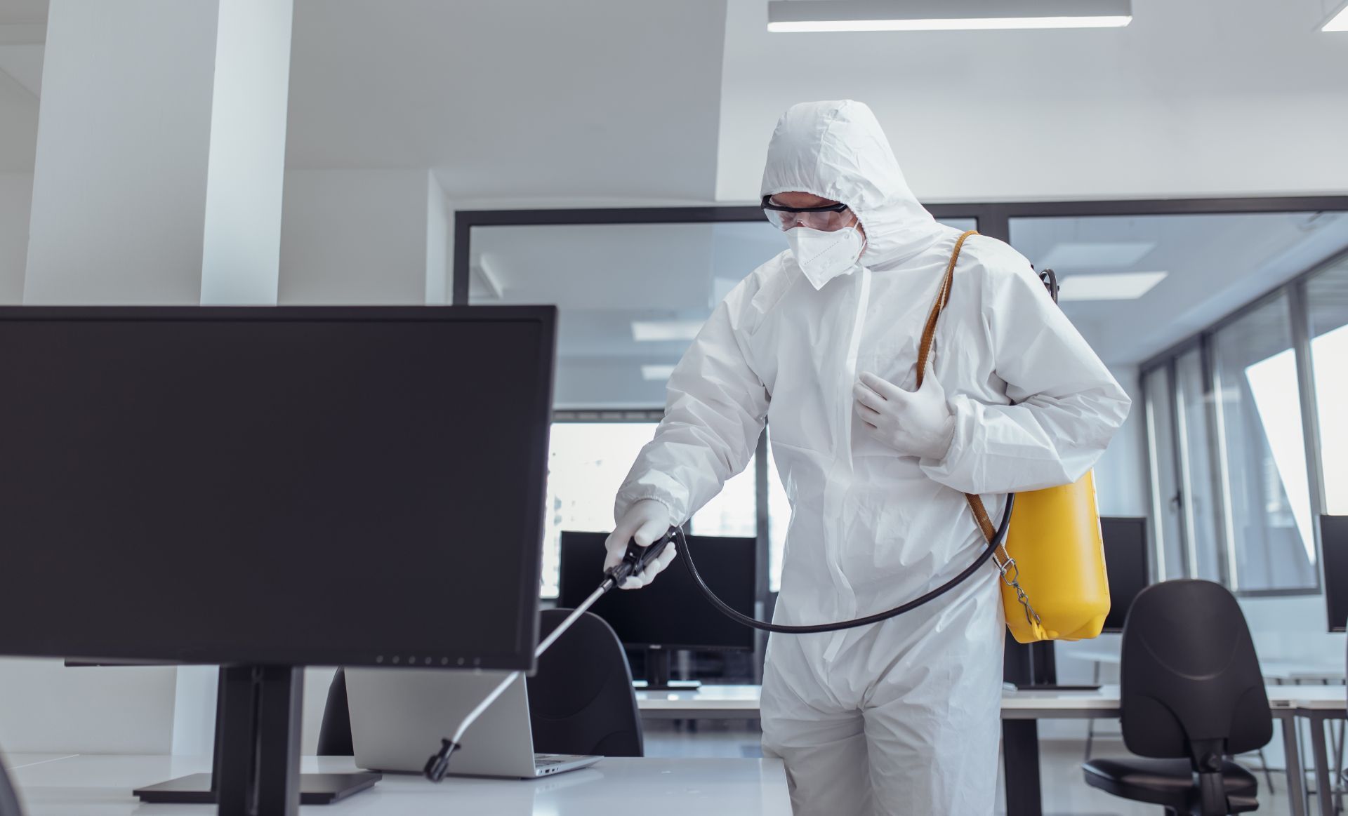 Worker in white protective suit spraying disinfectant in a bright room with a yellow tank sprayer