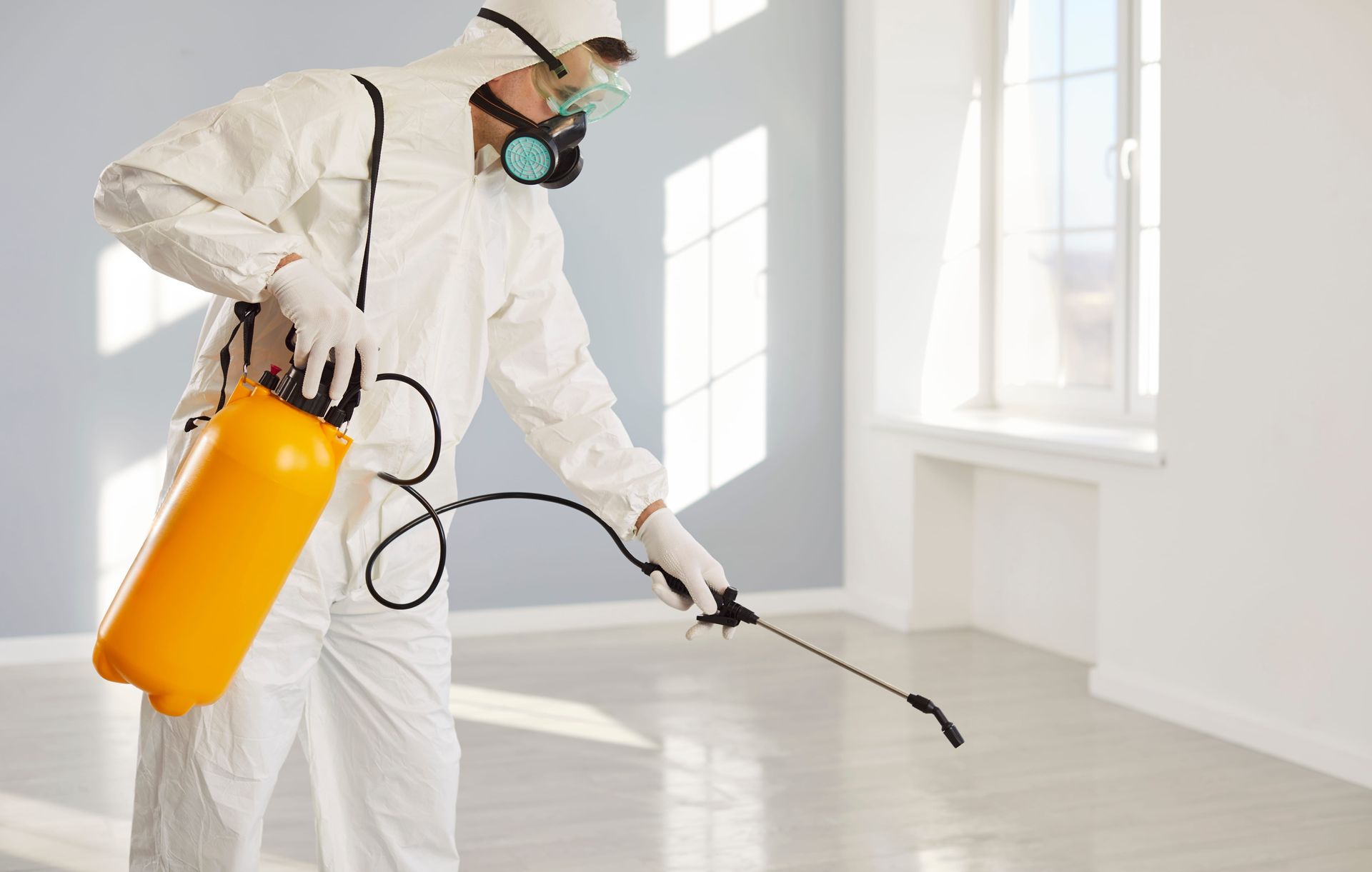Worker in white protective suit spraying disinfectant in a bright room with a yellow tank sprayer