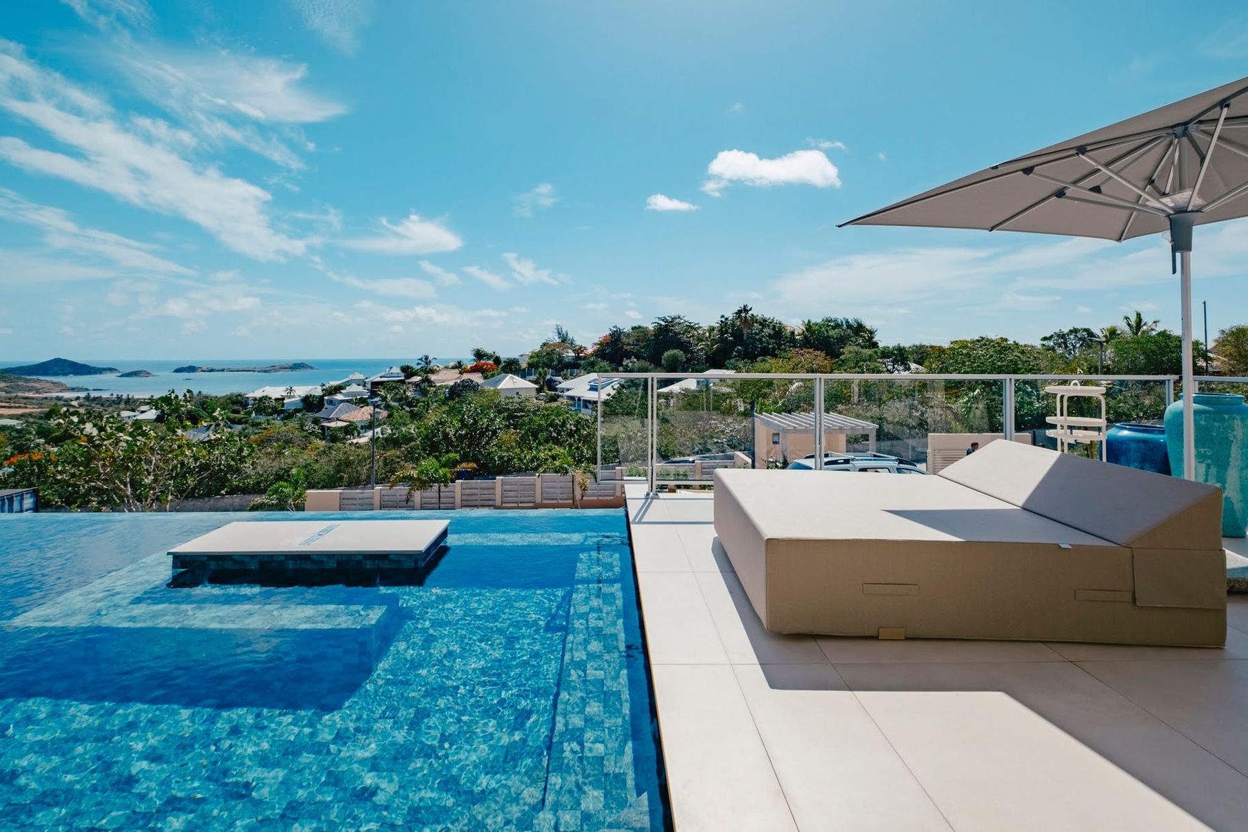 Modern white house with flat green roof, pool, and parked cars. Lush greenery surrounds the property.