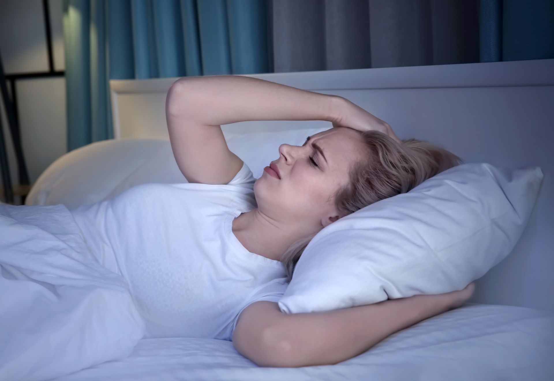 A person lies in bed in a dark room with a pained expression, hand held to their forehead as if experiencing a headache.
