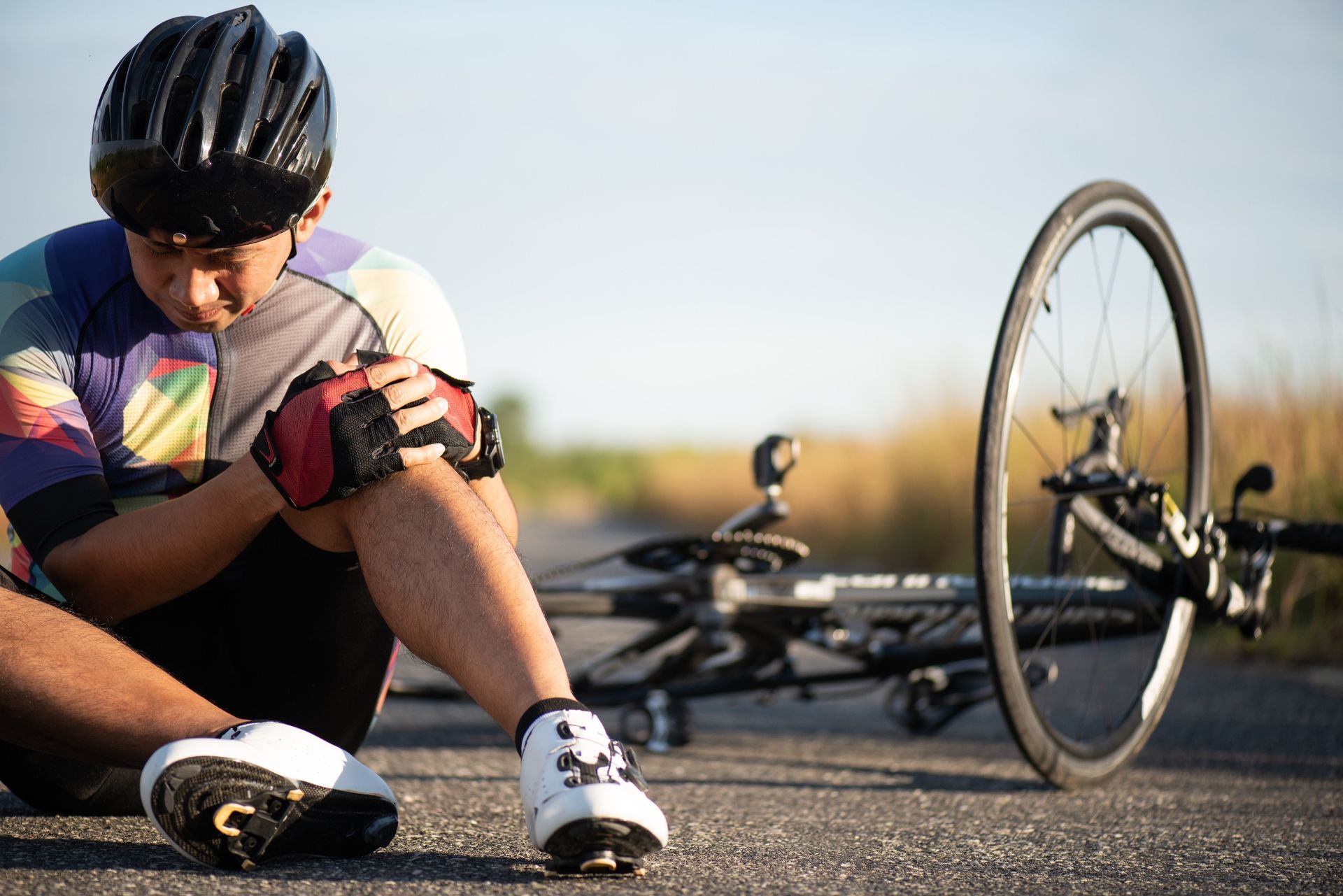 A cyclist wearing a helmet and gloves sits on the road in pain, holding their knee after a bike crash.