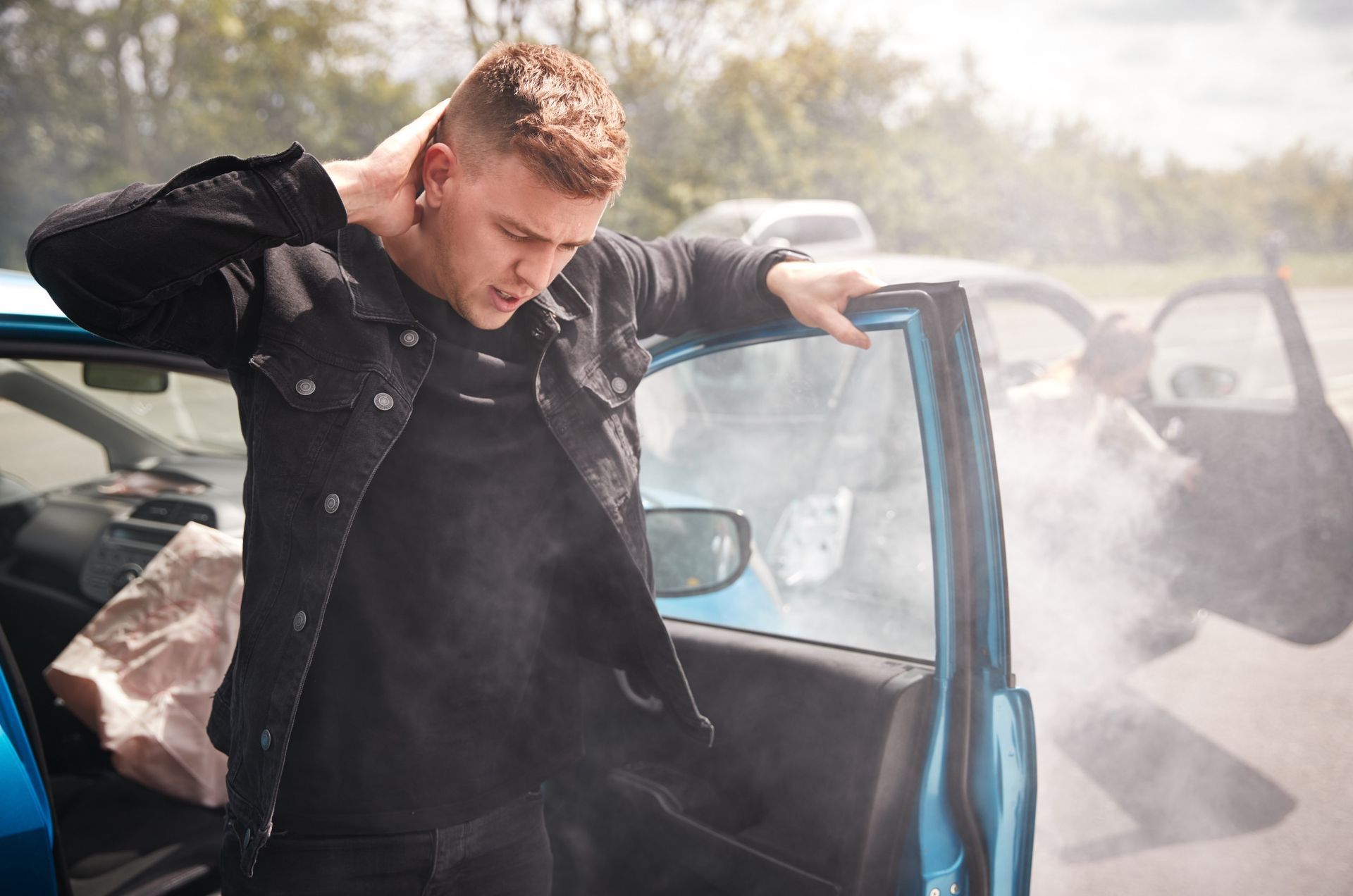 A person stands by a smoking, crashed blue car, holding their neck in pain after an accident.