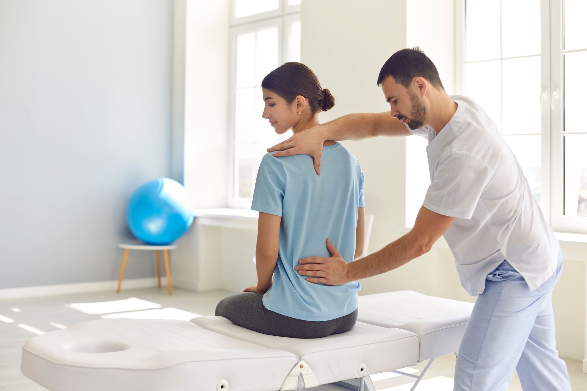 A physical therapist in white scrubs examines the back of a patient seated on a massage table in a bright clinic.