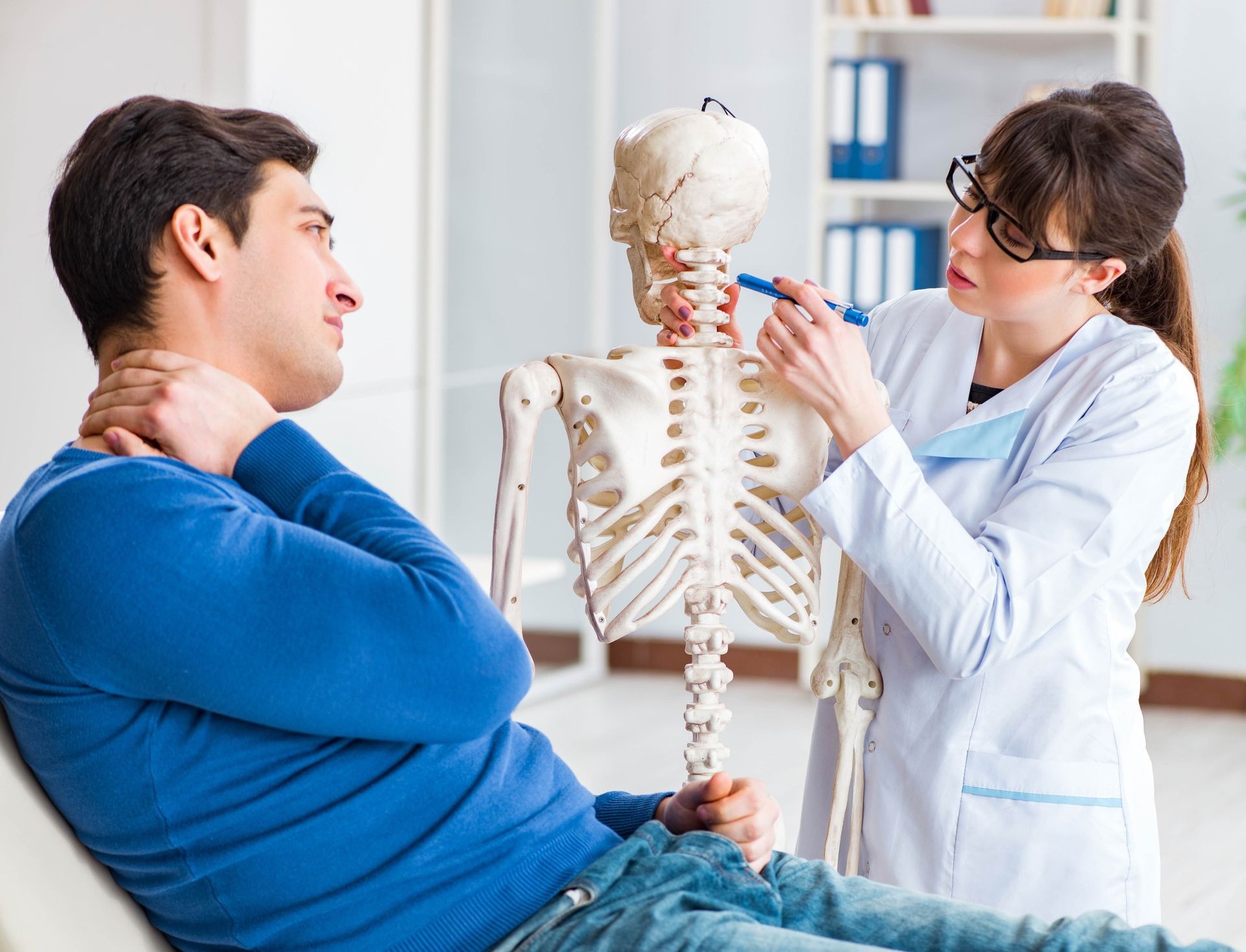 A healthcare professional uses a pen to point to the cervical spine on a skeletal model for a patient holding their neck.