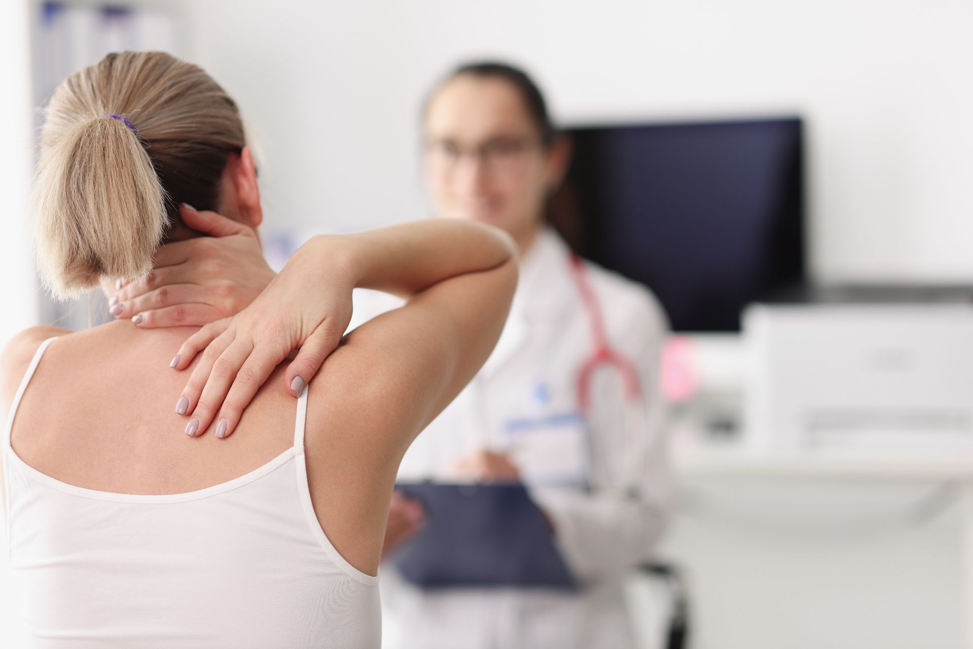 A person holding their neck in a doctor's office, with a medical professional in the blurred background.
