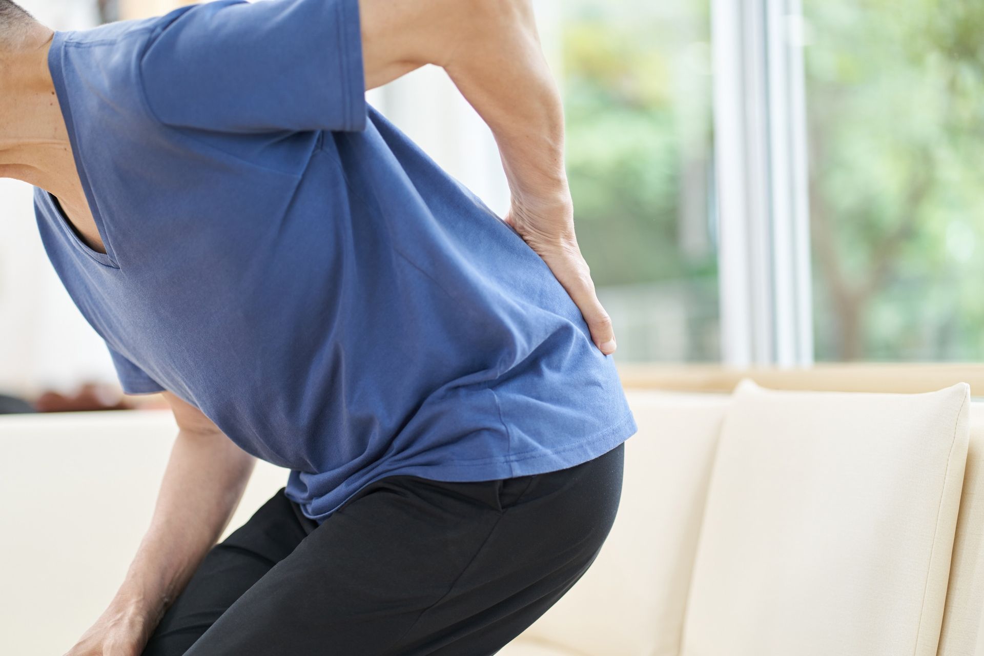 A person in a blue shirt leaning forward, holding their lower back due to pain in a brightly lit indoor setting.