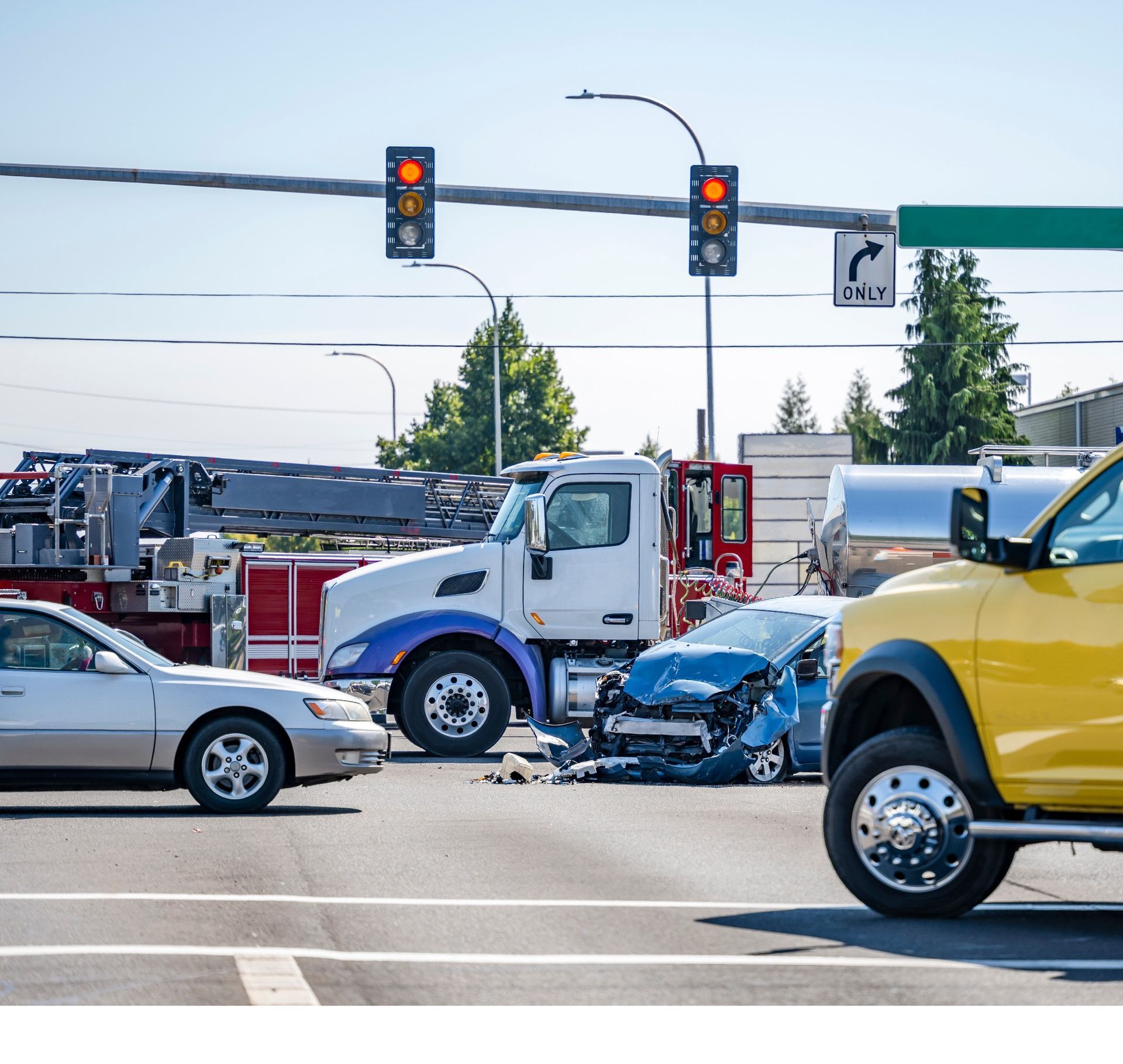 A car accident at an intersection featuring a blue car, a white semi-truck, a fire truck, and other vehicles.