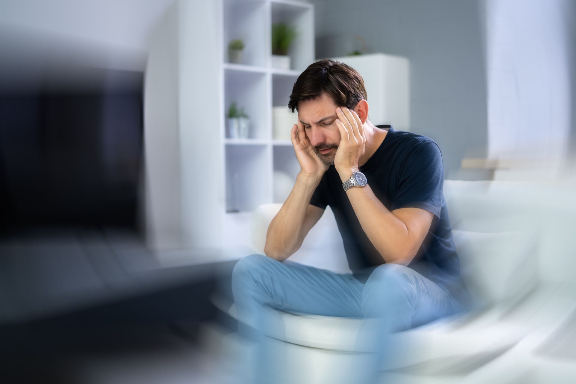 A person sits on a sofa, eyes closed, holding their temples with a pained expression, with a motion-blurred background.