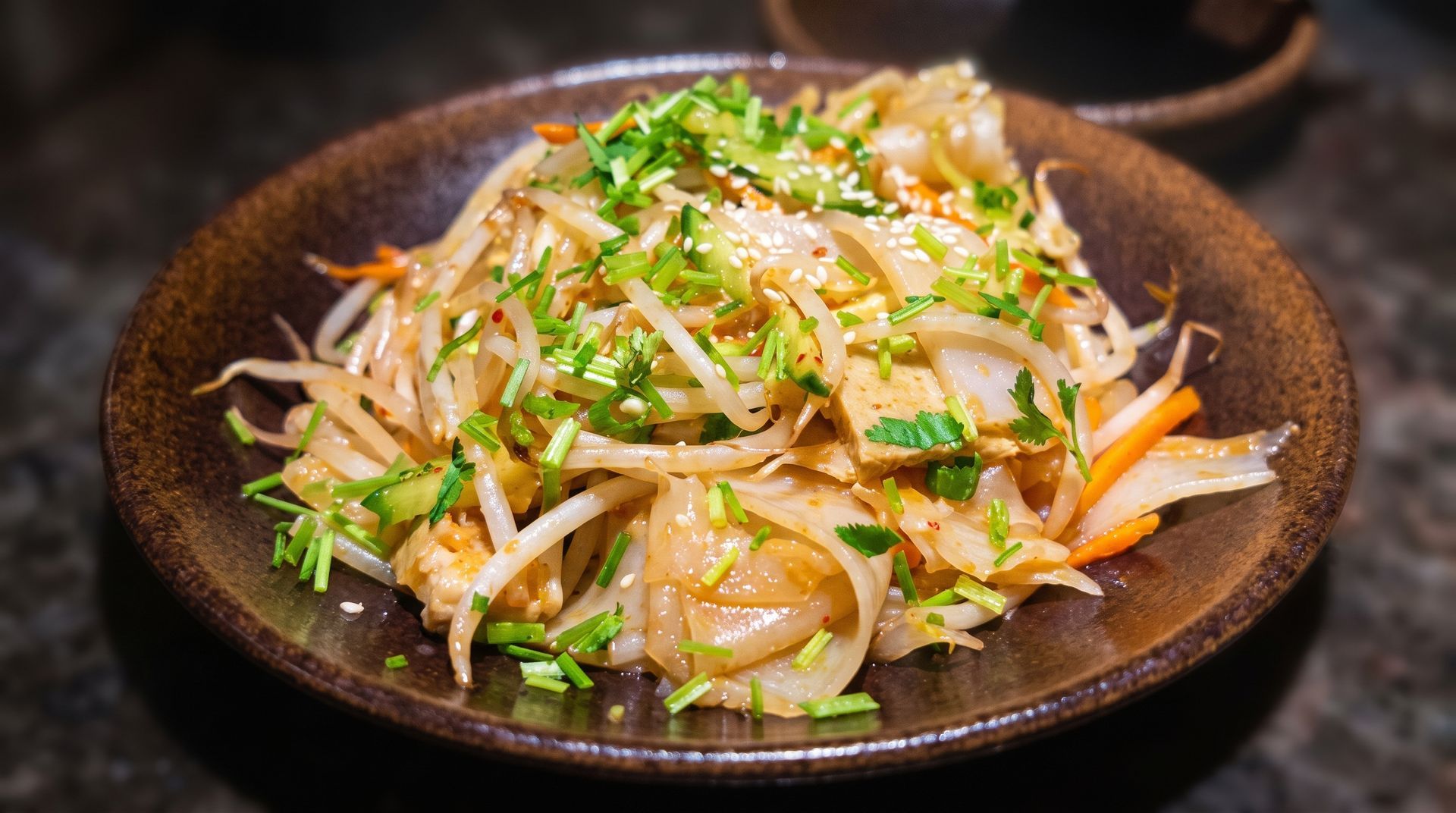 Stir-fried noodles with vegetables and herbs on a dark plate, garnished with sesame seeds.