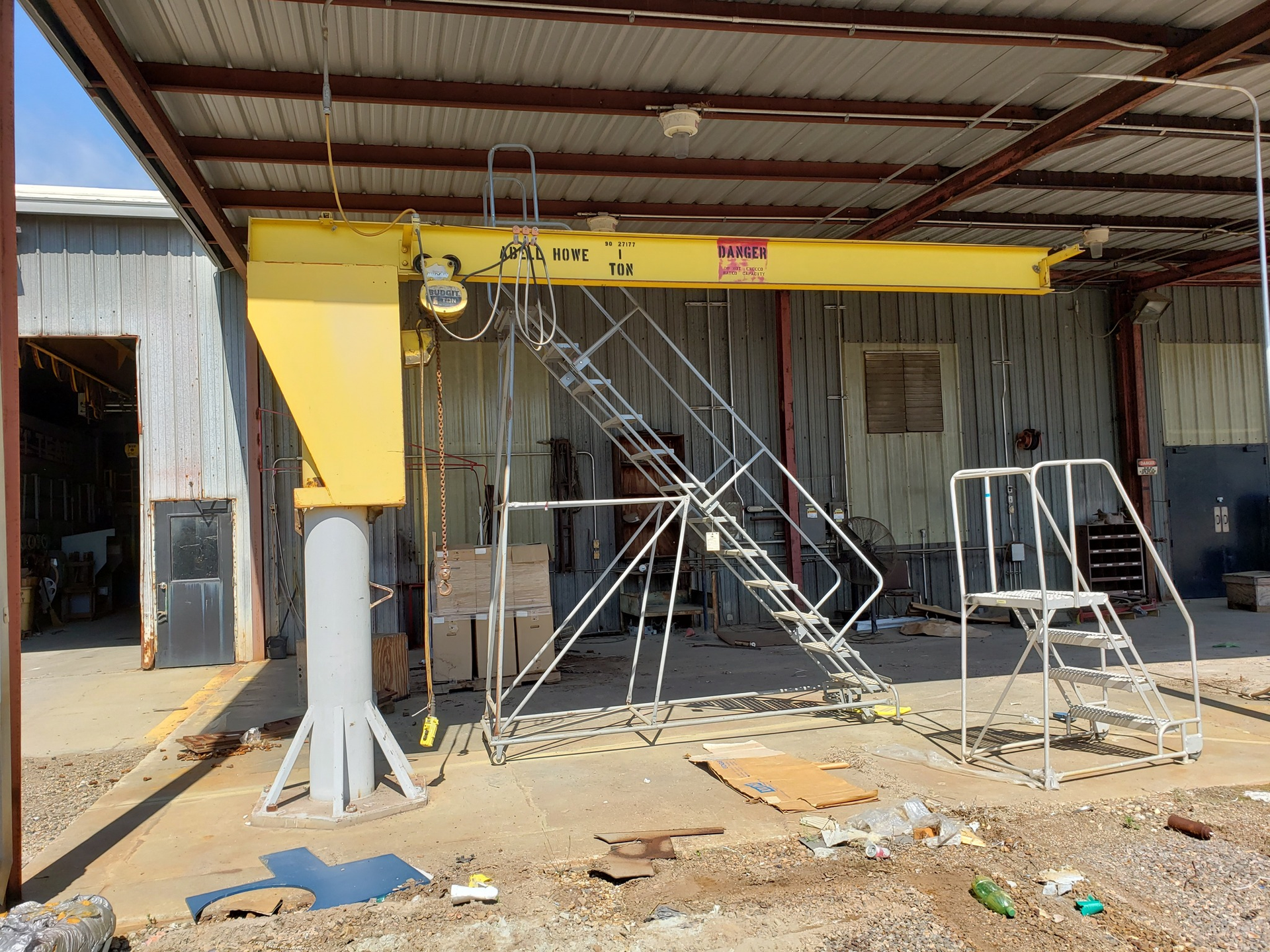 Yellow crane under a shelter, with metal stairs and various metal objects in a work area.