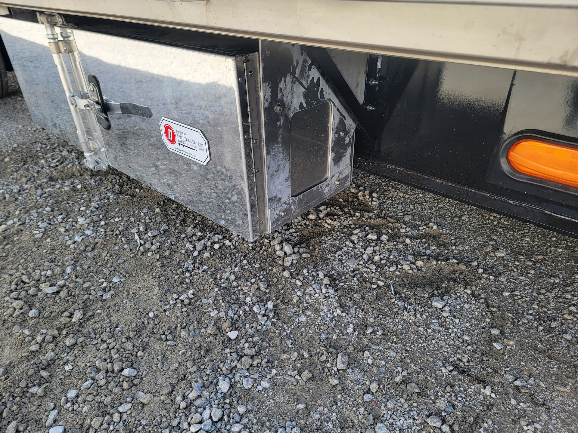 Silver metal storage box mounted on a black trailer, resting on gravel.