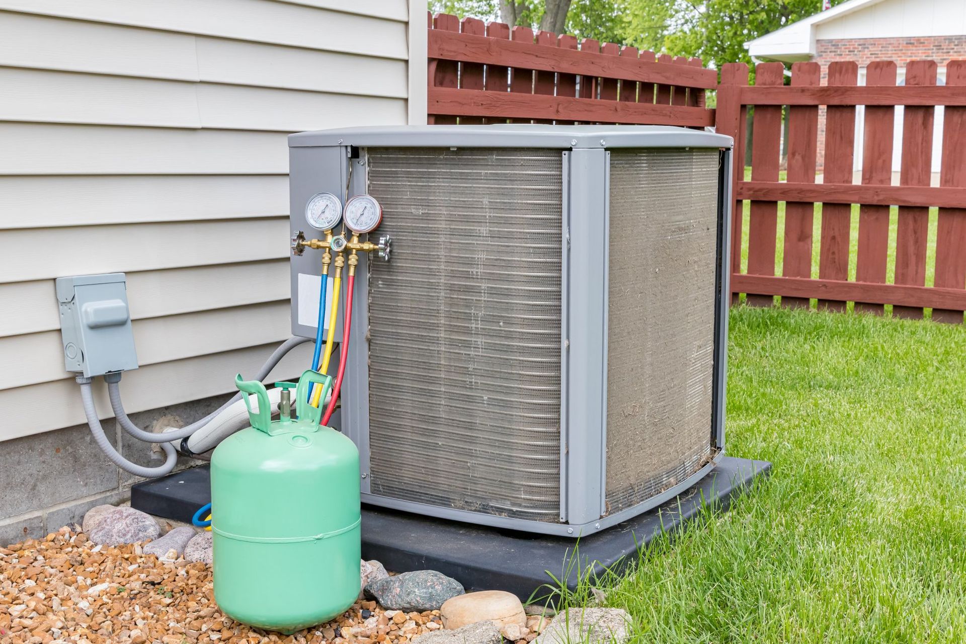Outdoor air conditioner unit beside a house with gauges, hoses, and a green refrigerant tank