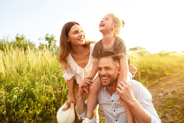 Family laughing outdoors in a sunny field, with a child on a man's shoulders and a woman beside them Family laughing outdoors in a sunny field, with a child on a man's shoulders and a woman beside them