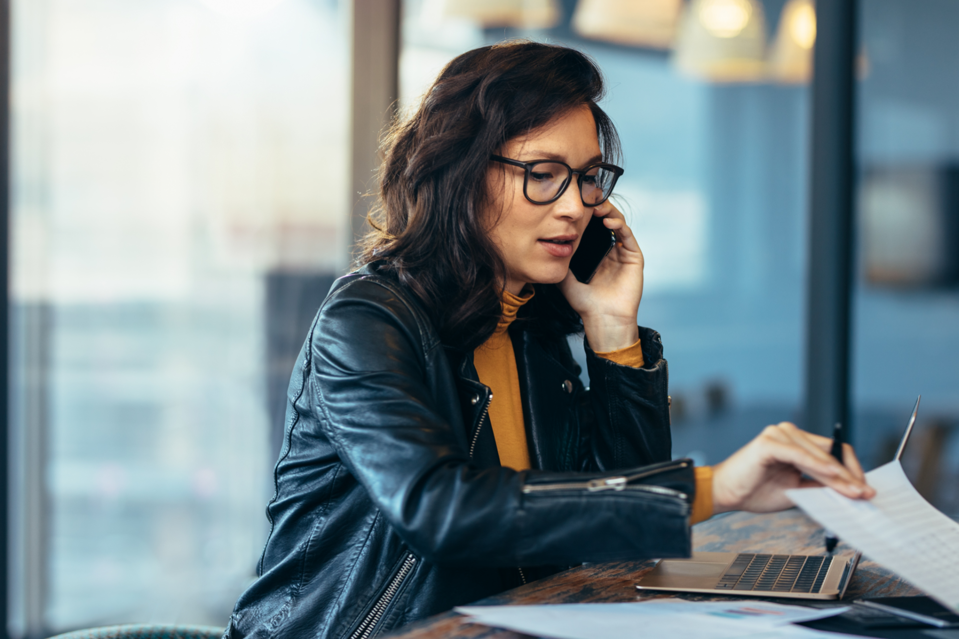 Woman in glasses and black jacket talking on a phone while reviewing papers at a laptop in an office