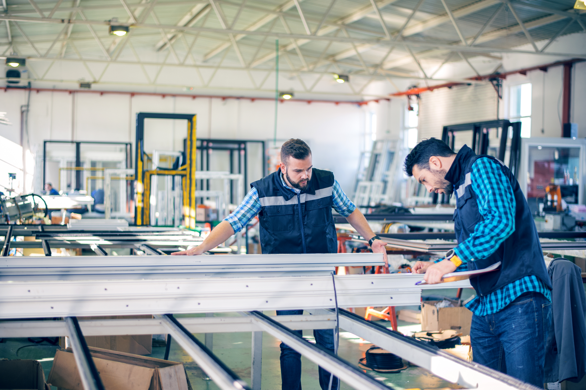 Two workers measure materials in a bright industrial workshop with large machinery and worktables.
