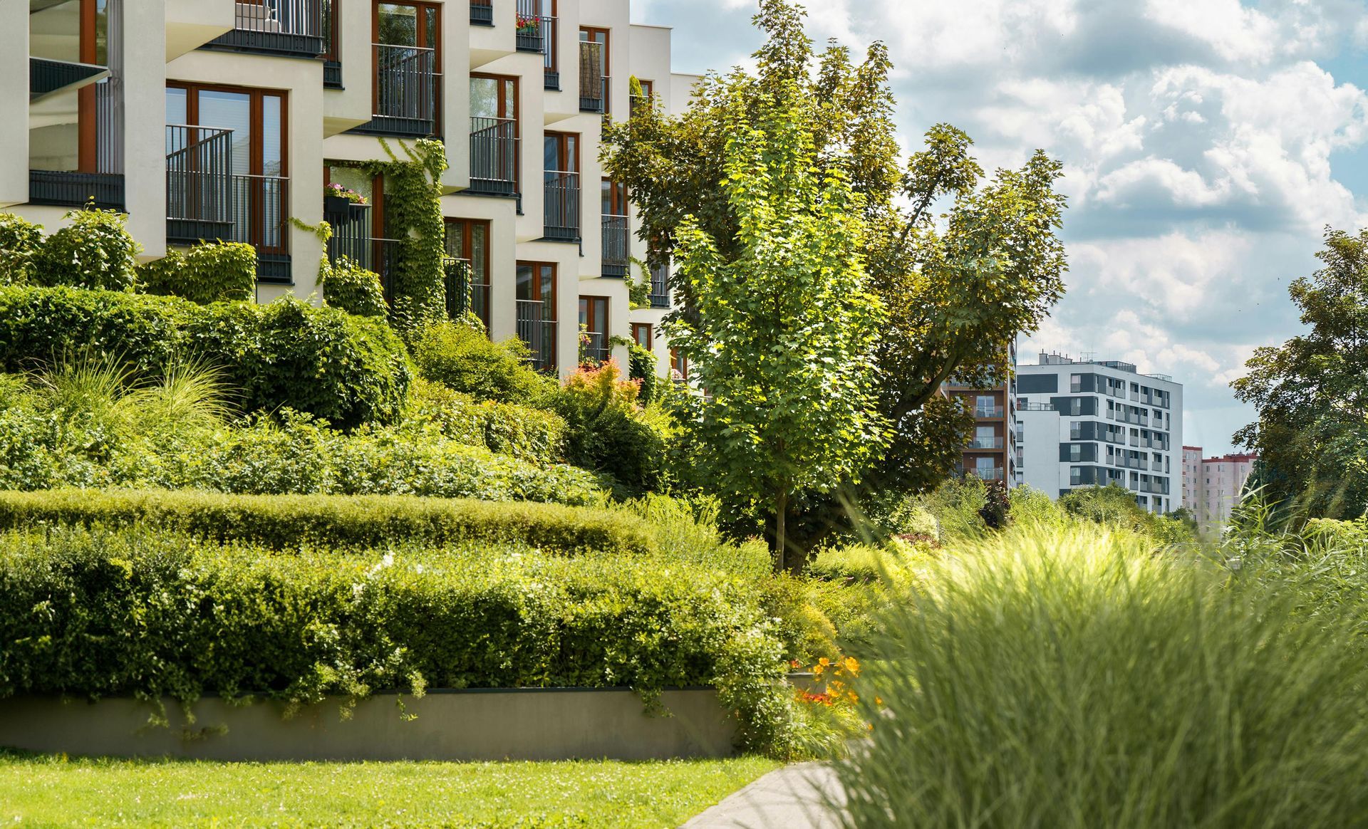Modern apartment buildings behind lush green landscaping and tall grasses under a partly cloudy sky