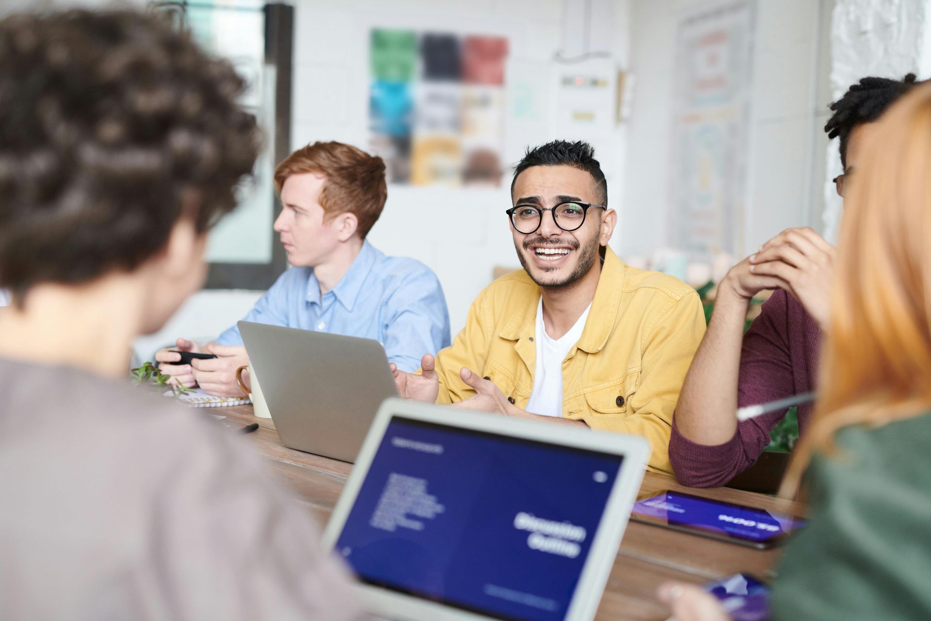 Colleagues meeting around a table with laptops, one smiling and holding a notebook in a bright office