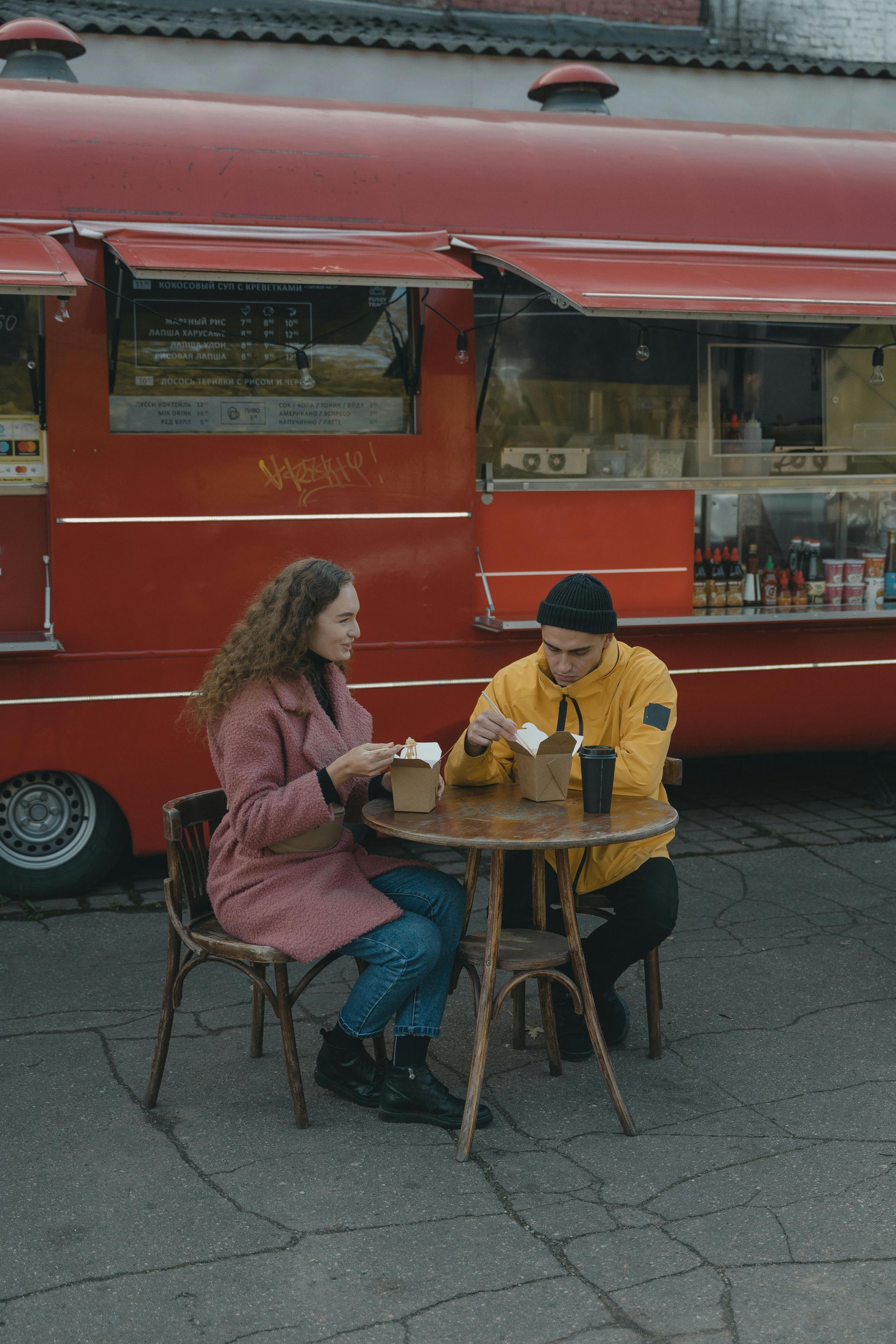 Two people sit at a small table outside a red food truck, eating and talking.