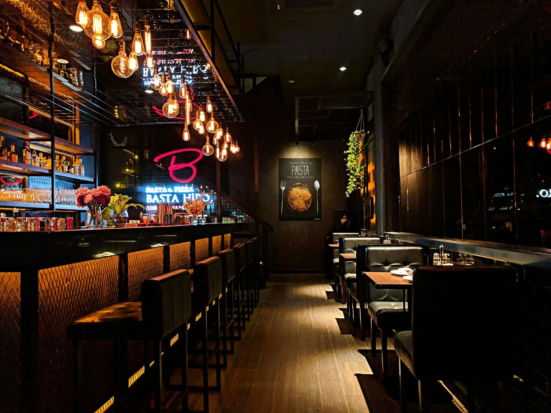 Dimly lit modern bar interior with neon sign, hanging lights, and empty stools along the counter.