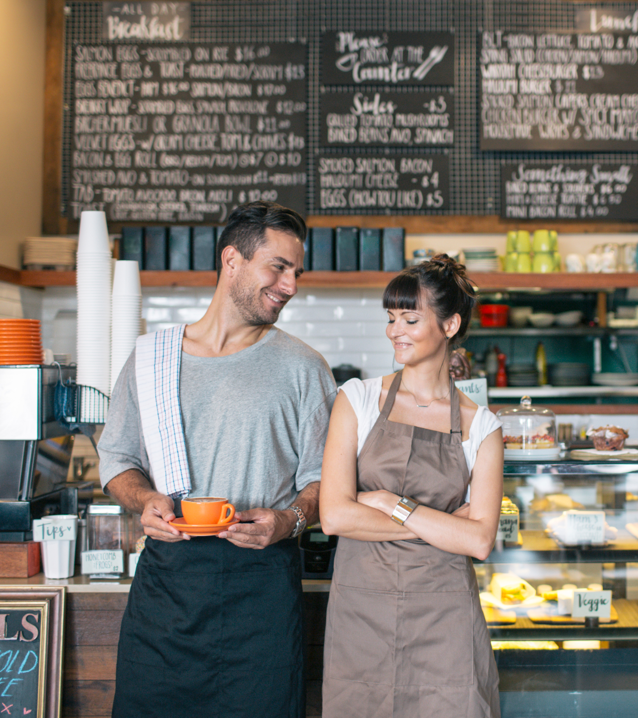 Two café workers in aprons smiling at each other behind the counter with menu boards in the background