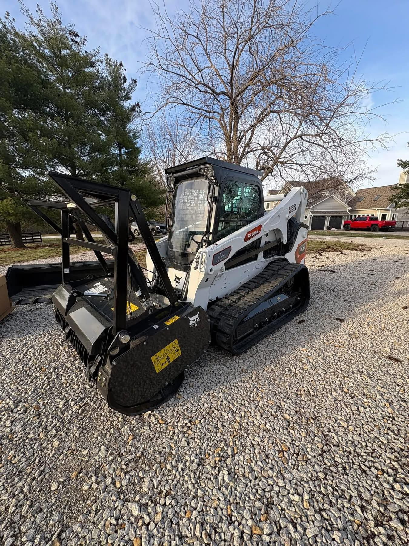 White skid-steer loader with black attachment parked on gravel beside a shed and trees.