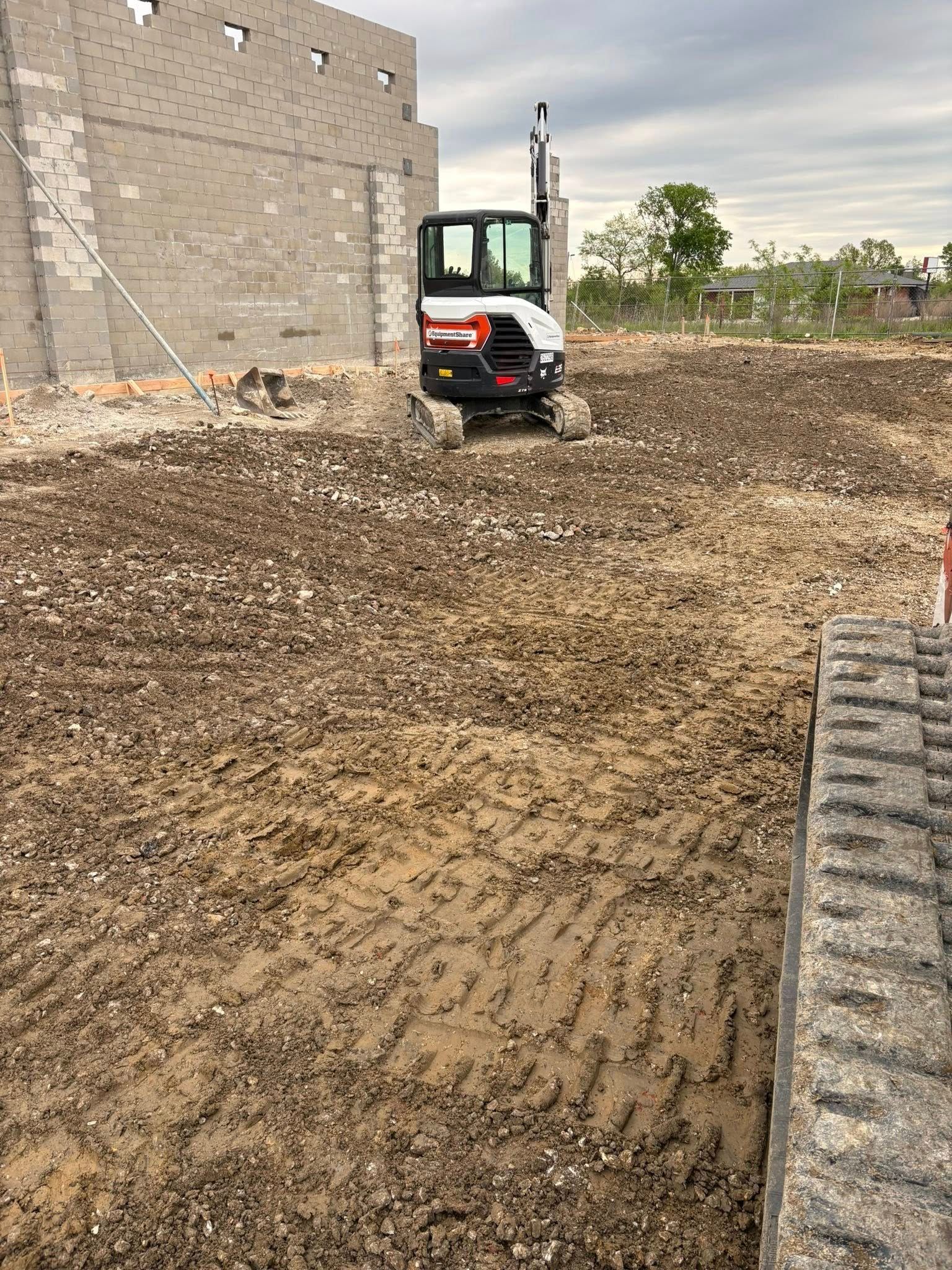 Excavator grading a muddy construction site beside a brick wall and a large tire track in the foreground