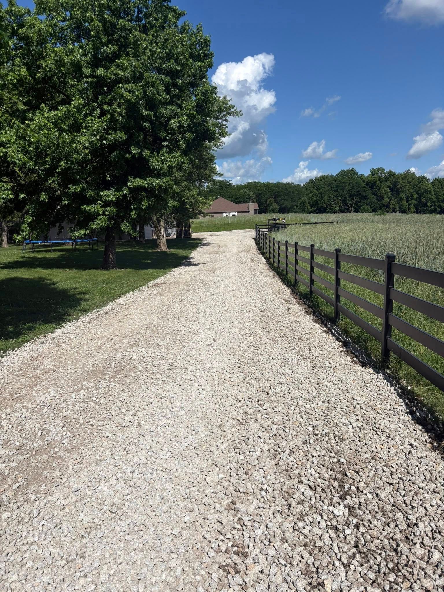 Gravel driveway beside a black fence and green field, leading toward a house under a blue sky.