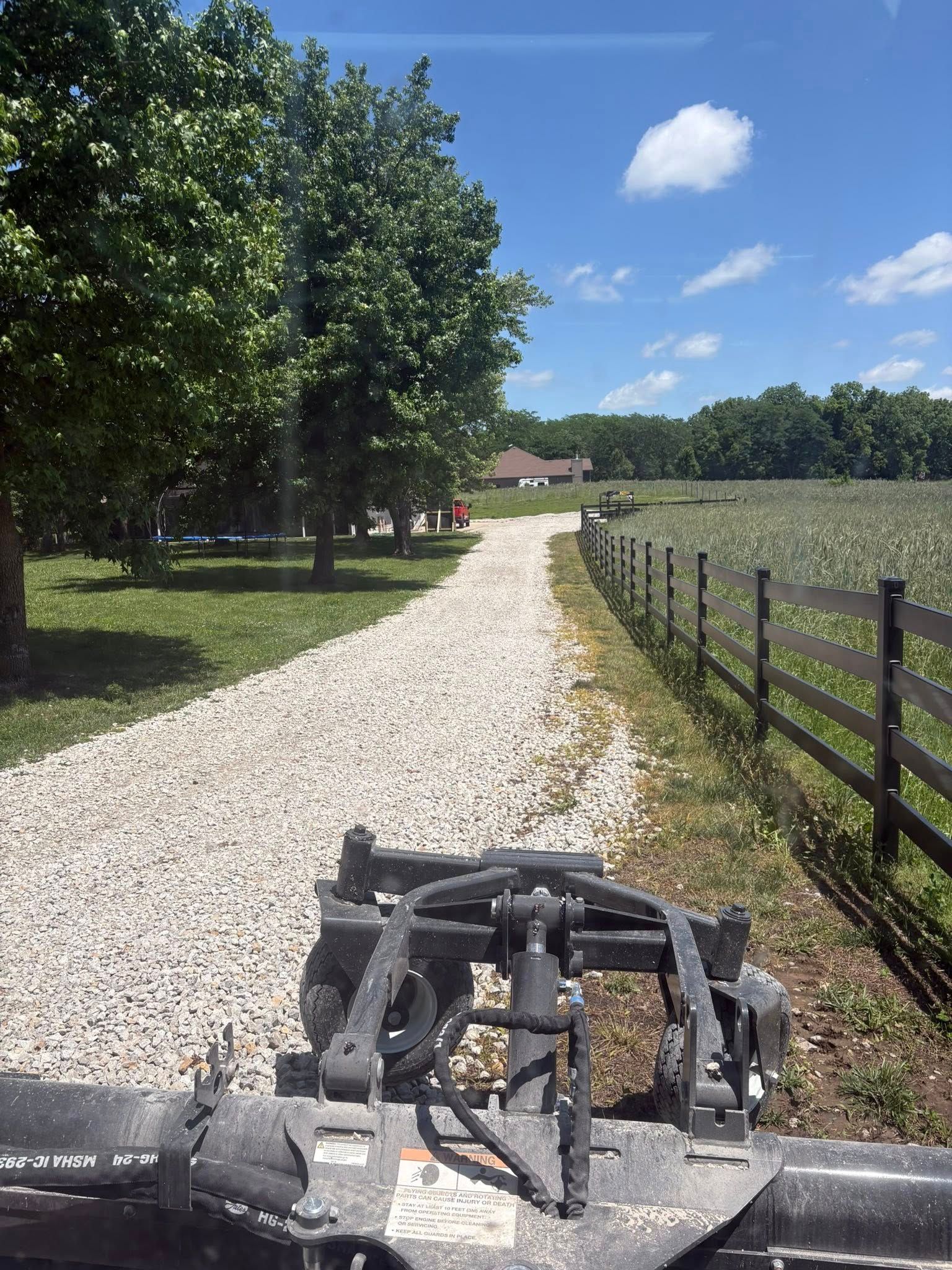 Gravel farm lane ahead with trees on left, fenced field on right, and a mower deck at the bottom foreground