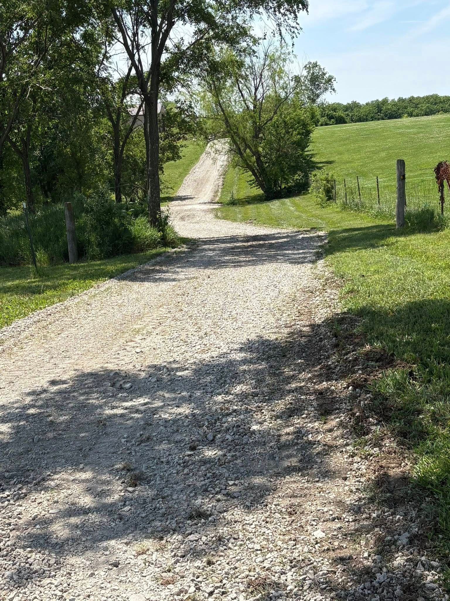 Gravel path winding through a sunny green field with trees and fence posts
