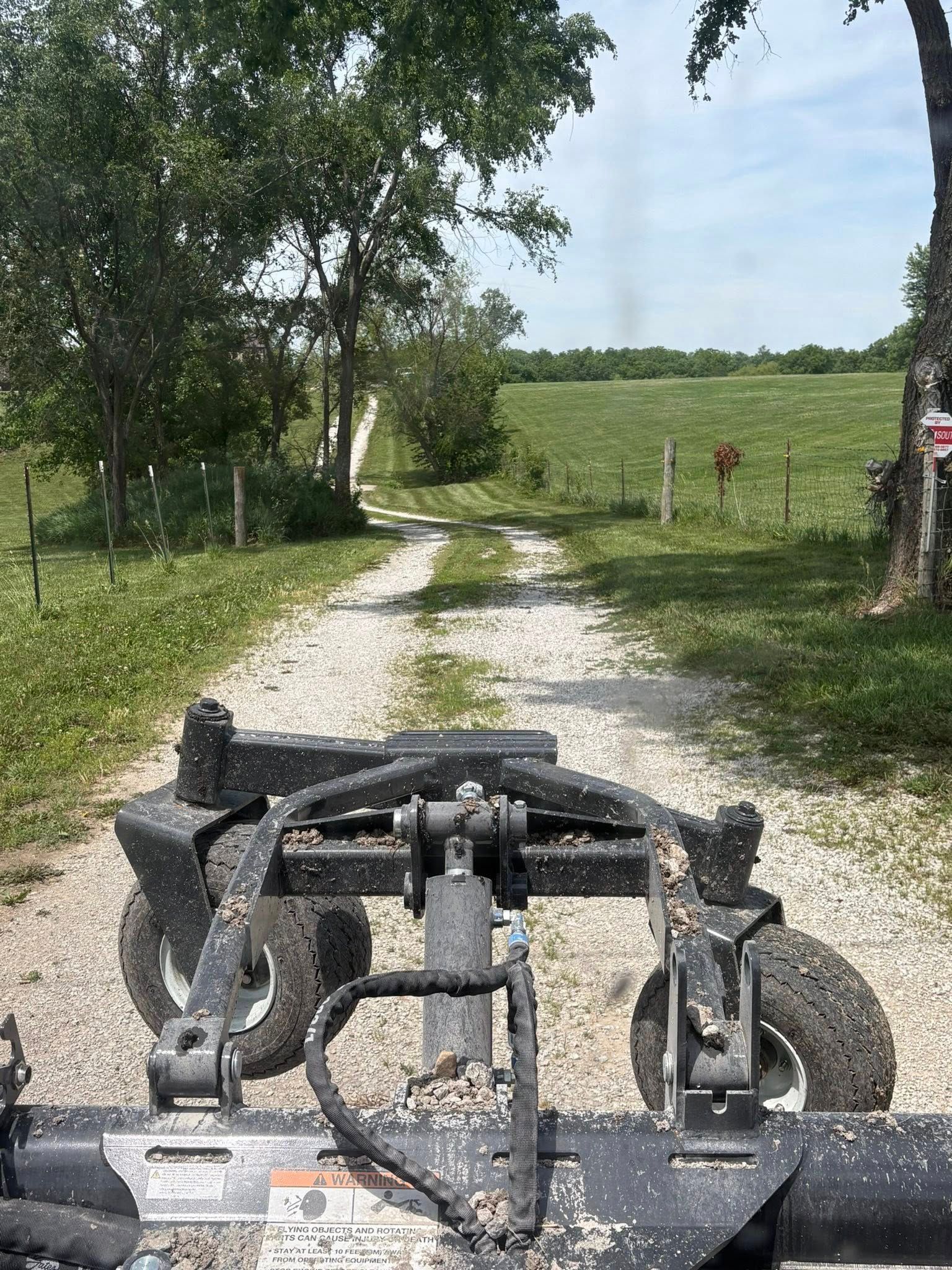 Mud-covered ATV on a gravel path beside a grassy field and trees under a bright sky