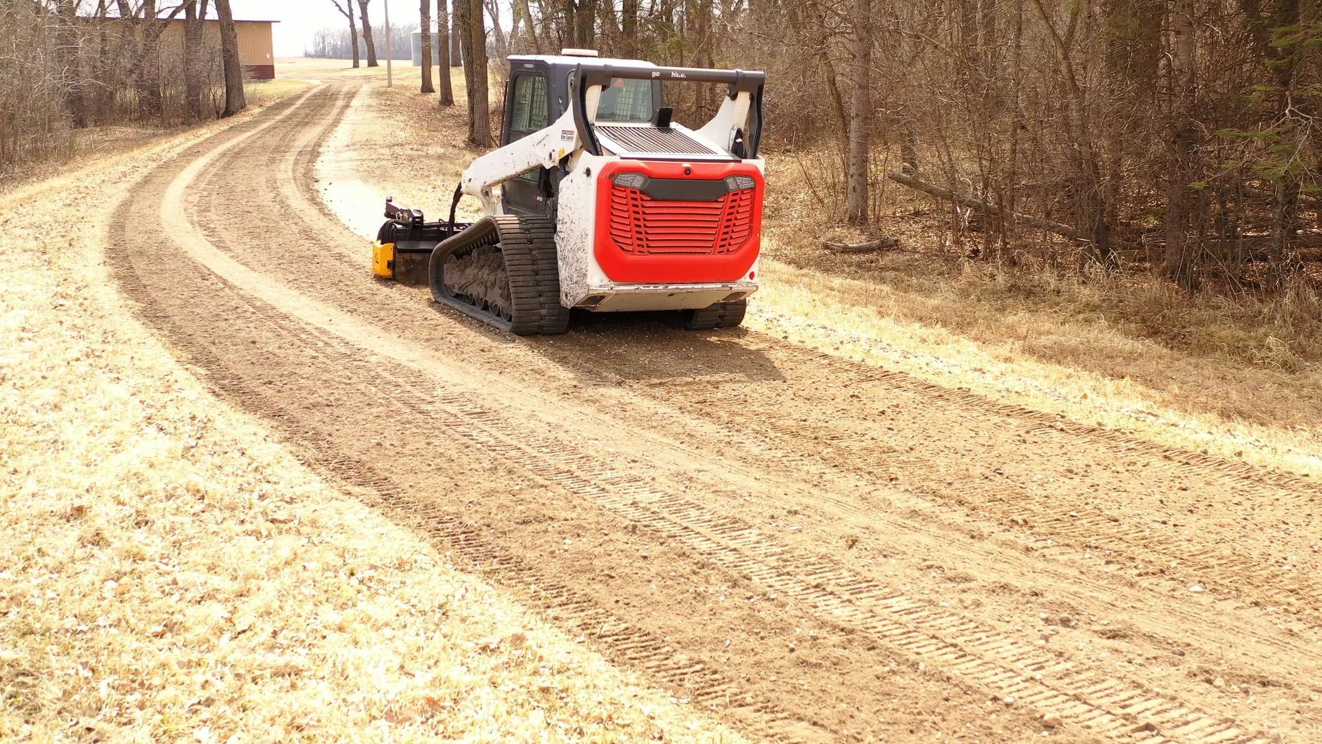 Compact skid steer clearing a curved dirt road, leaving fresh tracks in a wooded area.