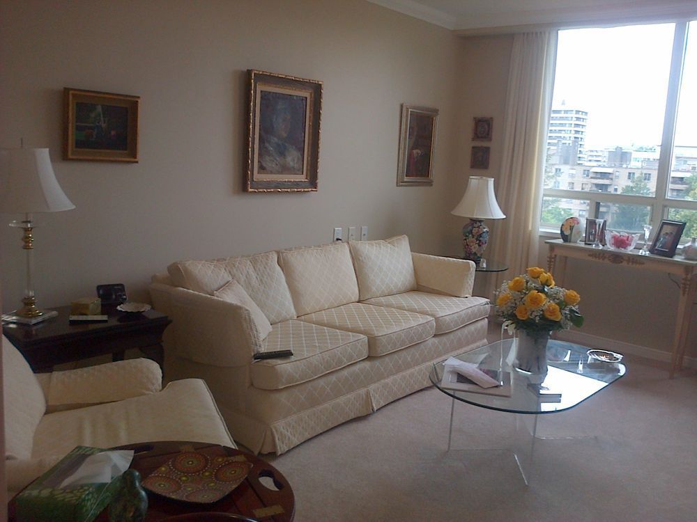 Beige living room with sofa, chair, paintings, glass coffee table, and window overlooking a city.