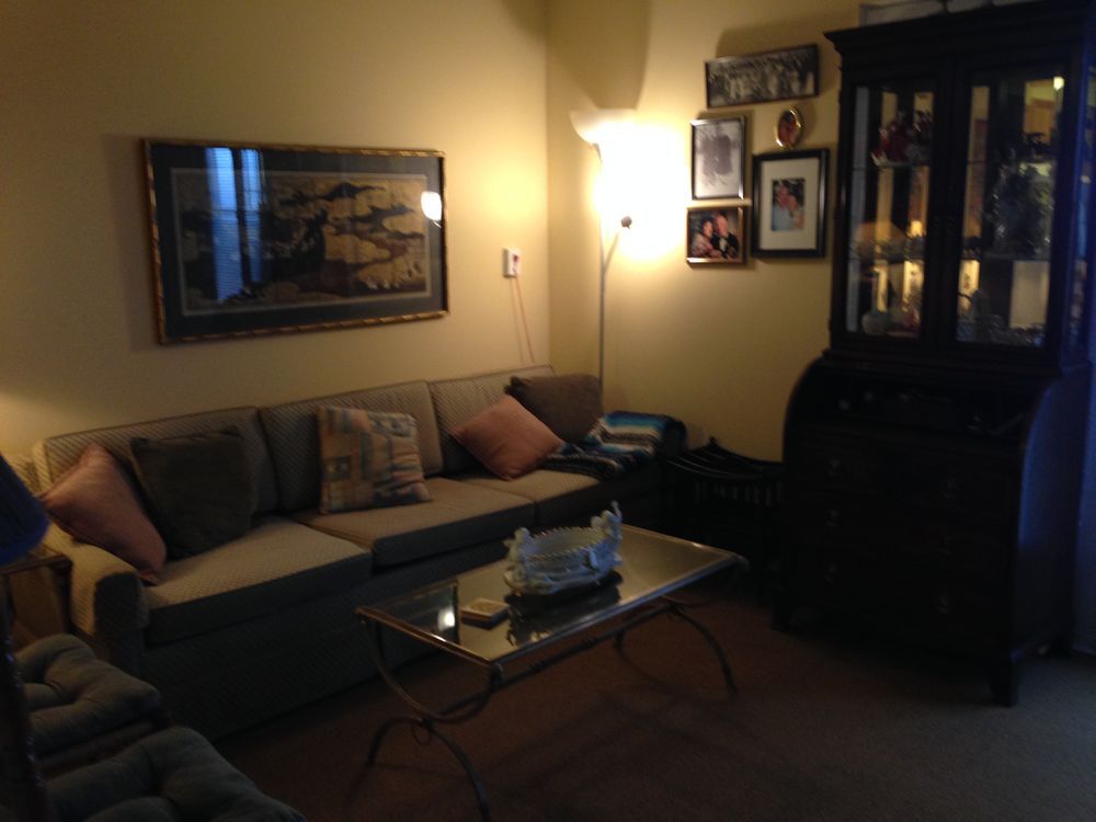 Living room with sofa, coffee table, and dark cabinet. Light from a floor lamp illuminates the space.