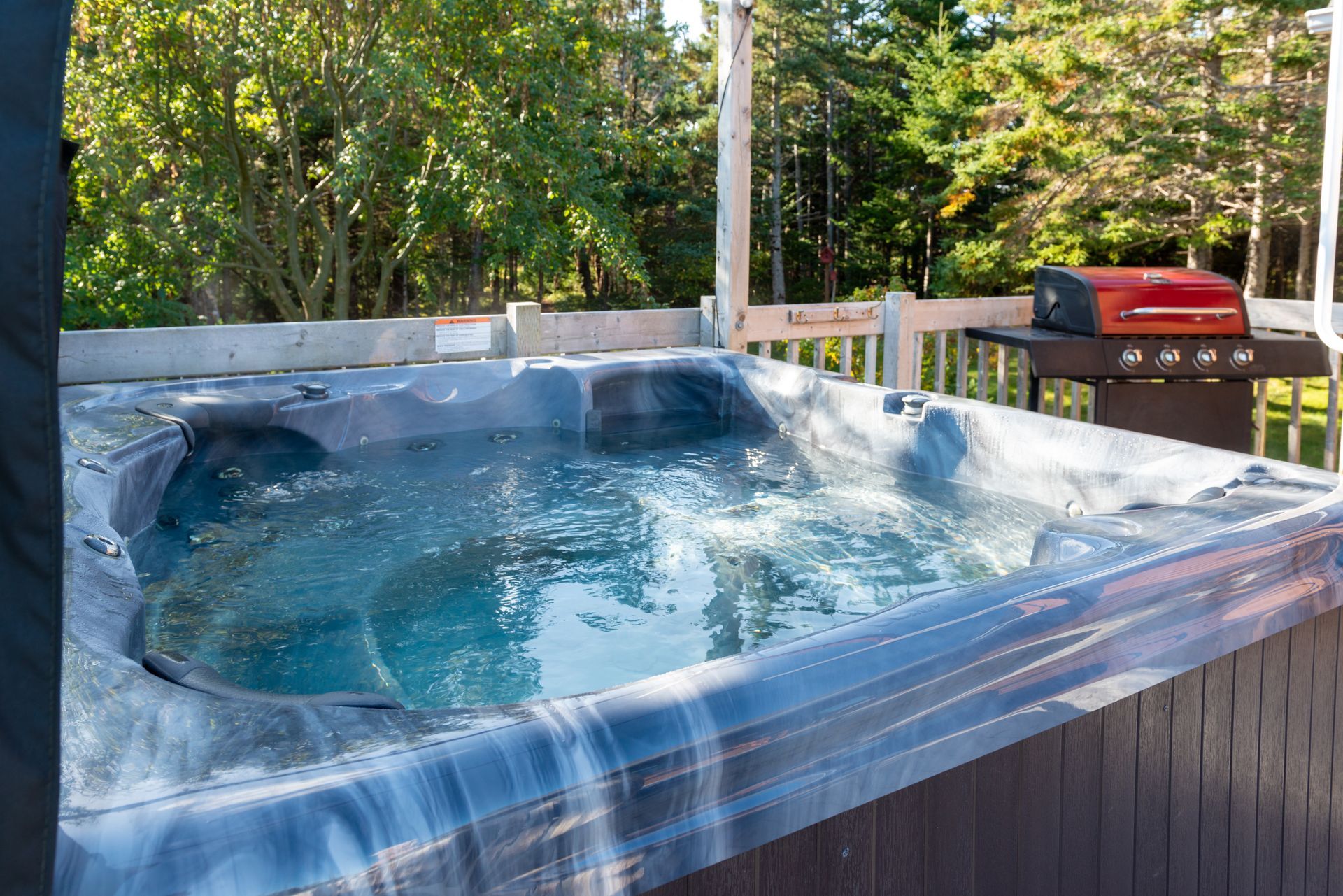 Hot tub on a deck with water bubbling, grill in background, trees in the distance.