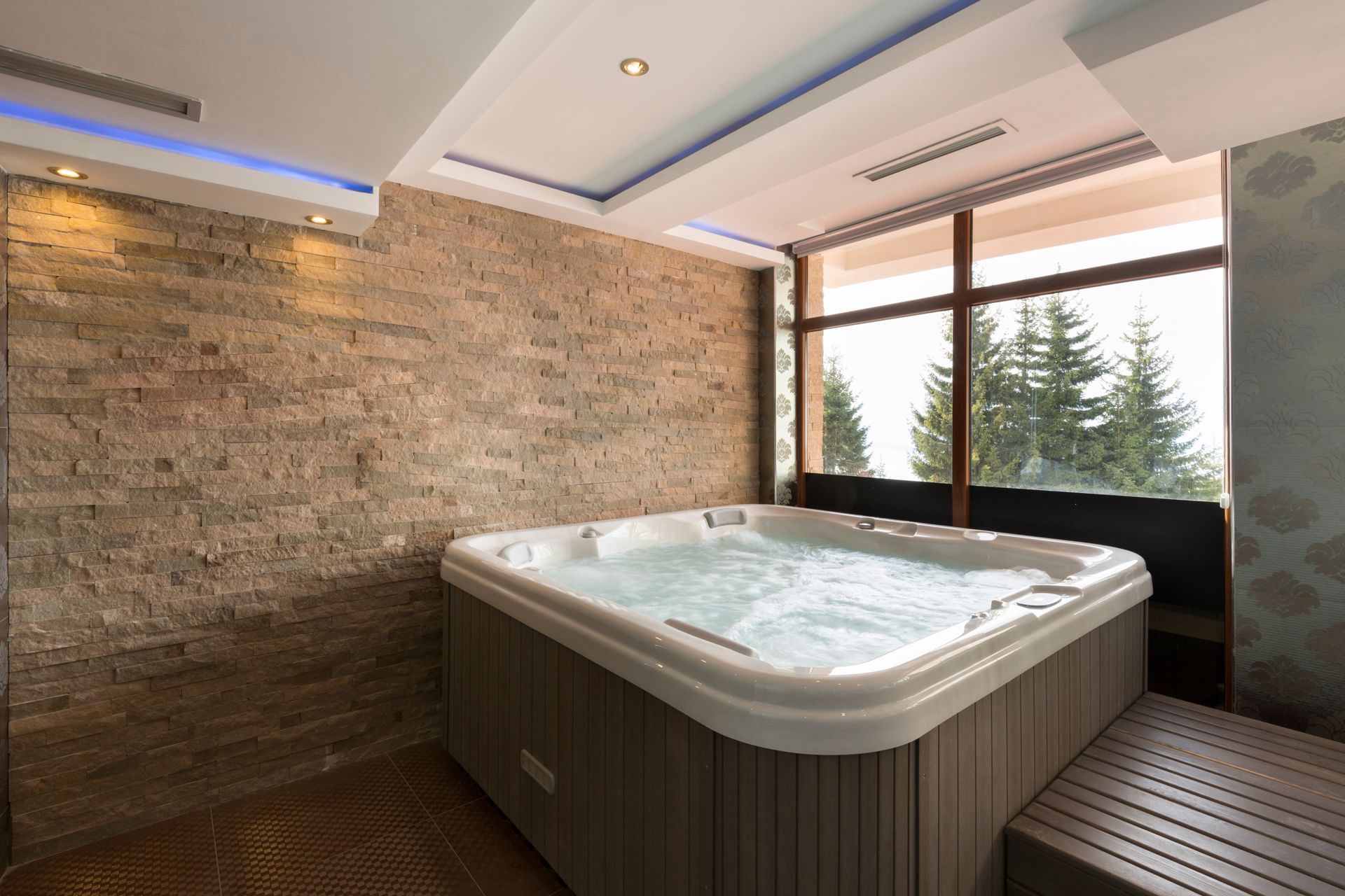 A hot tub with bubbly water is indoors next to a large window overlooking trees.