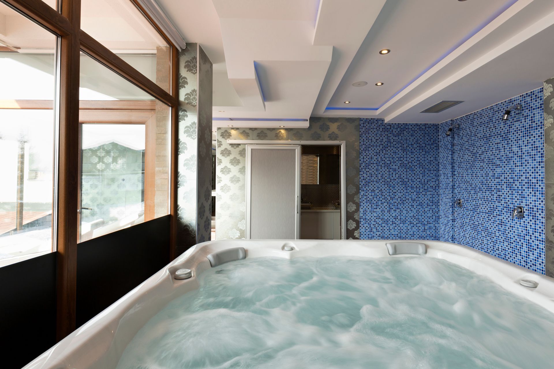 Whirlpool spa in a room with a blue tiled wall, a window, and a white ceiling with recessed lights.