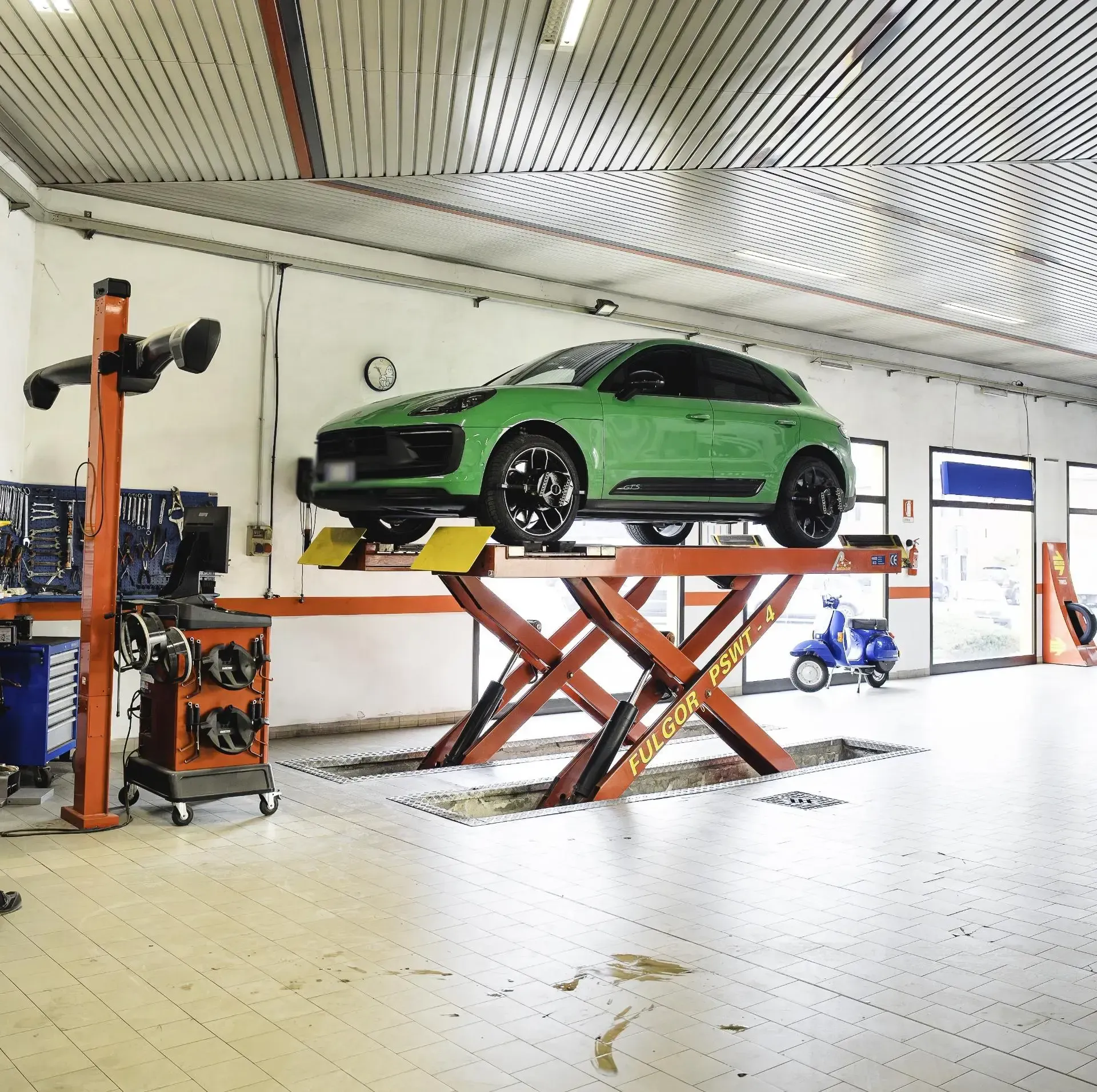 Un'auto verde brillante sollevata su un ponte idraulico a forbice all'interno di un'officina meccanica ben illuminata con pavimento piastrellato.