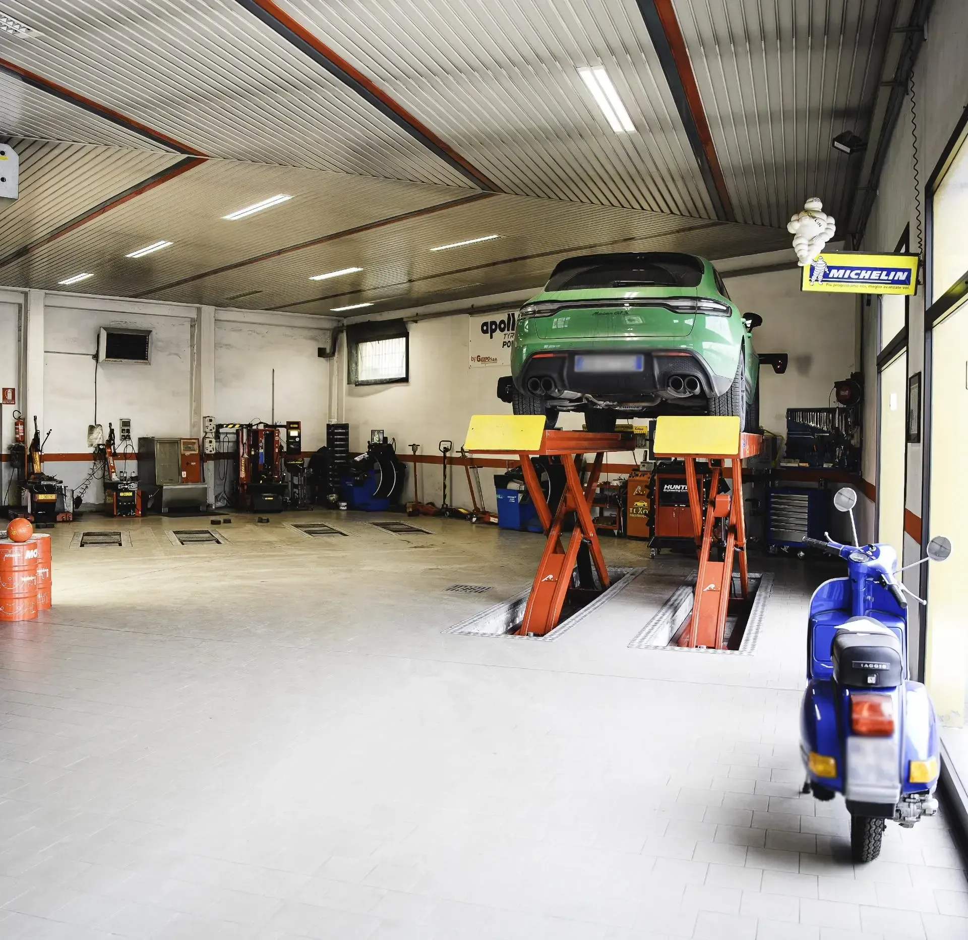 Un'officina meccanica luminosa con un'auto verde sollevata su un ponte idraulico e uno scooter blu parcheggiato sul pavimento di cemento.
