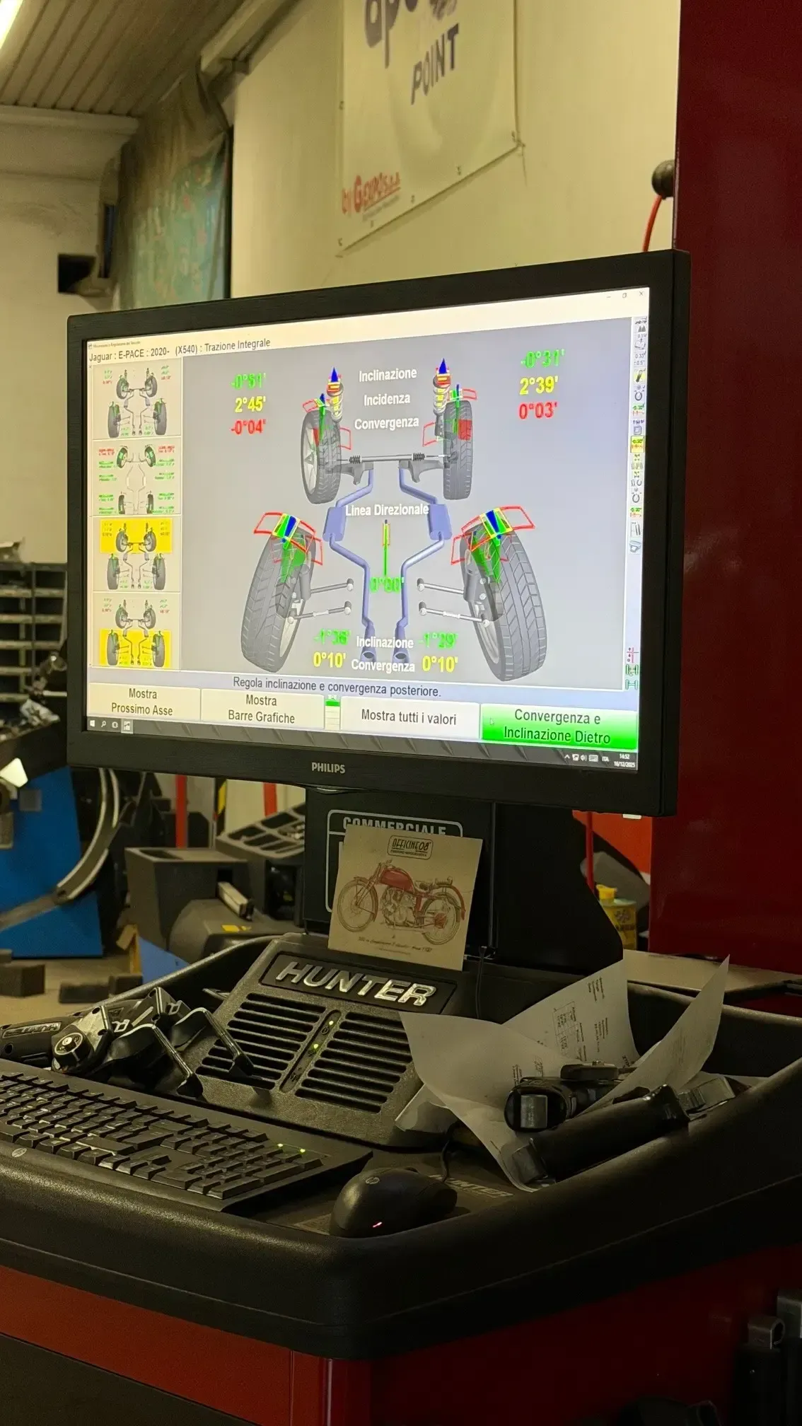 In un'officina meccanica, un monitor mostra una schermata diagnostica dell'allineamento delle ruote con diverse misurazioni evidenziate in rosso.