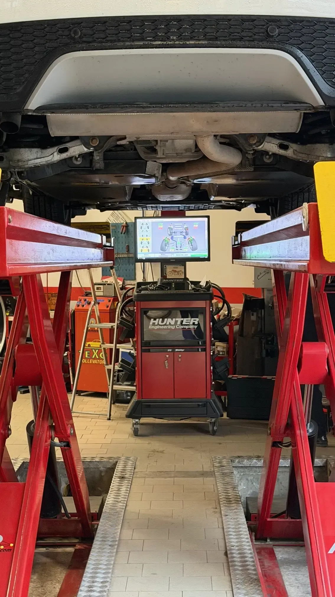 Un veicolo su un sollevatore idraulico rosso all'interno di un'officina meccanica, con al centro una macchina computerizzata per l'allineamento delle ruote.