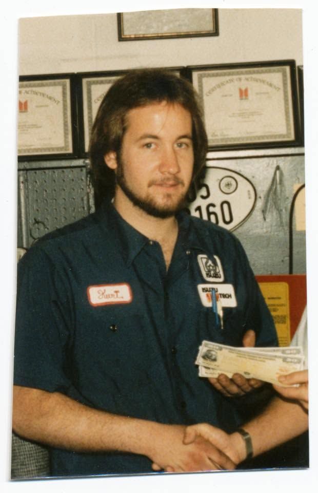 Man in navy work shirt holding papers, standing indoors in front of framed certificates and signs | Certified Import Repair