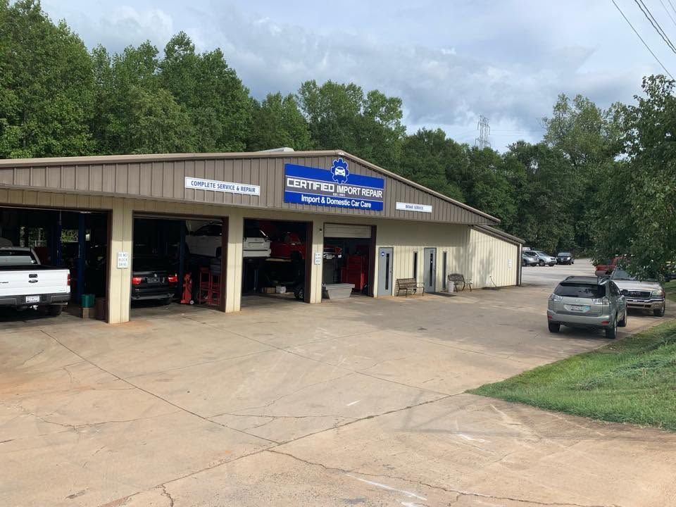 Auto repair shop with open bays, parked vehicles, and a blue sign under a cloudy sky | Certified Import Repair