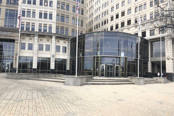 Glass entrance to a modern building with flags on poles, set in a paved plaza.