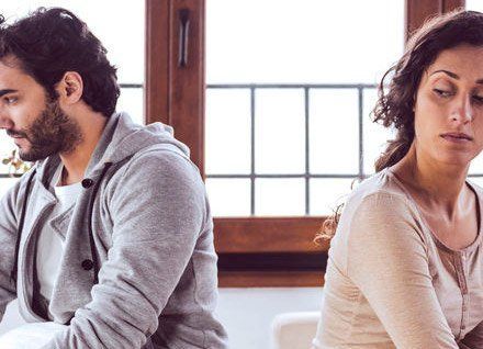 Man and woman, seated back-to-back, look somber. Interior setting, natural light.